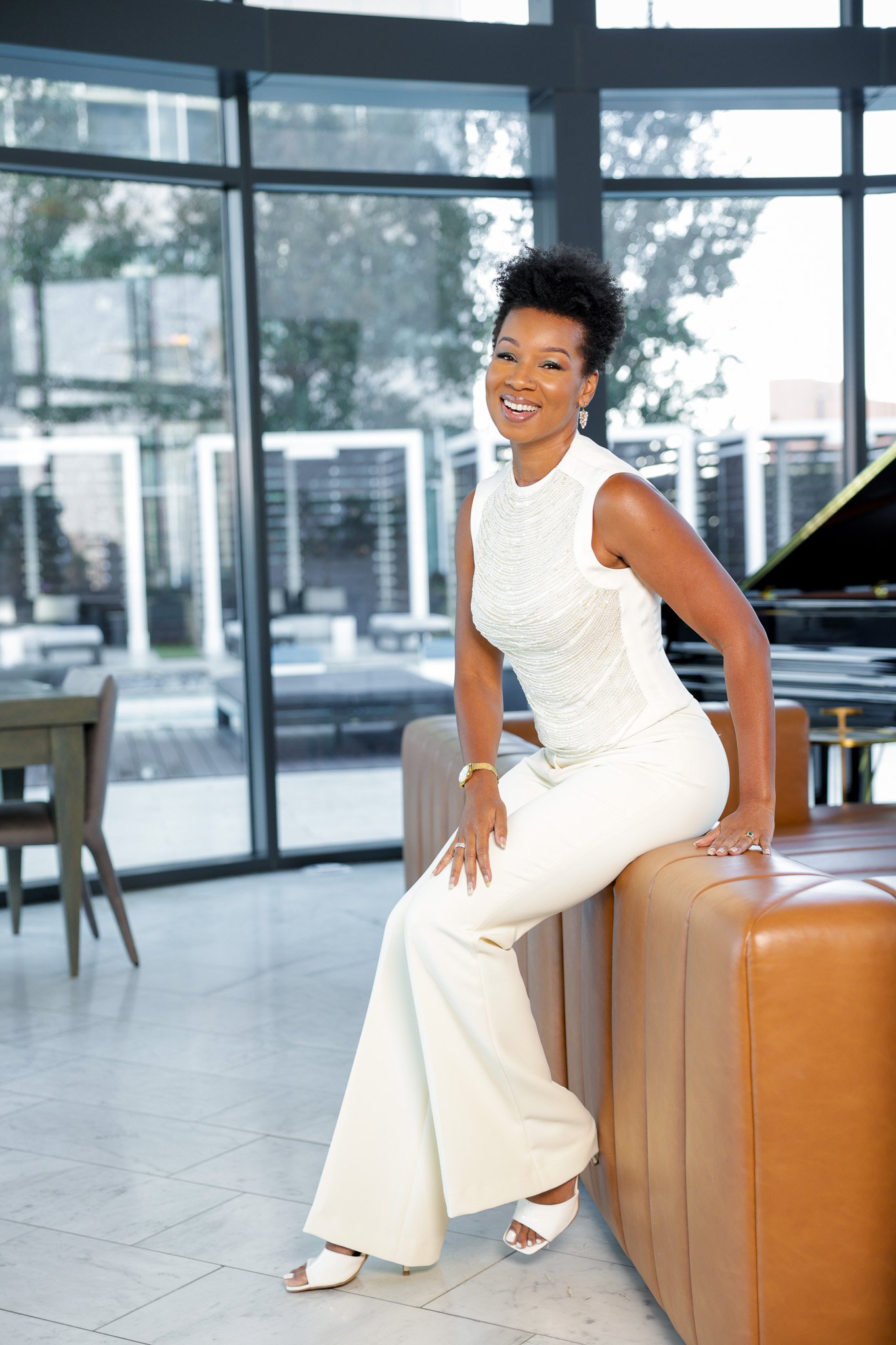 A woman in an elegant white dress and heels sitting on the edge of a tan leather sofa, smiling in a modern indoor setting with large glass windows and outdoor trees in the background.