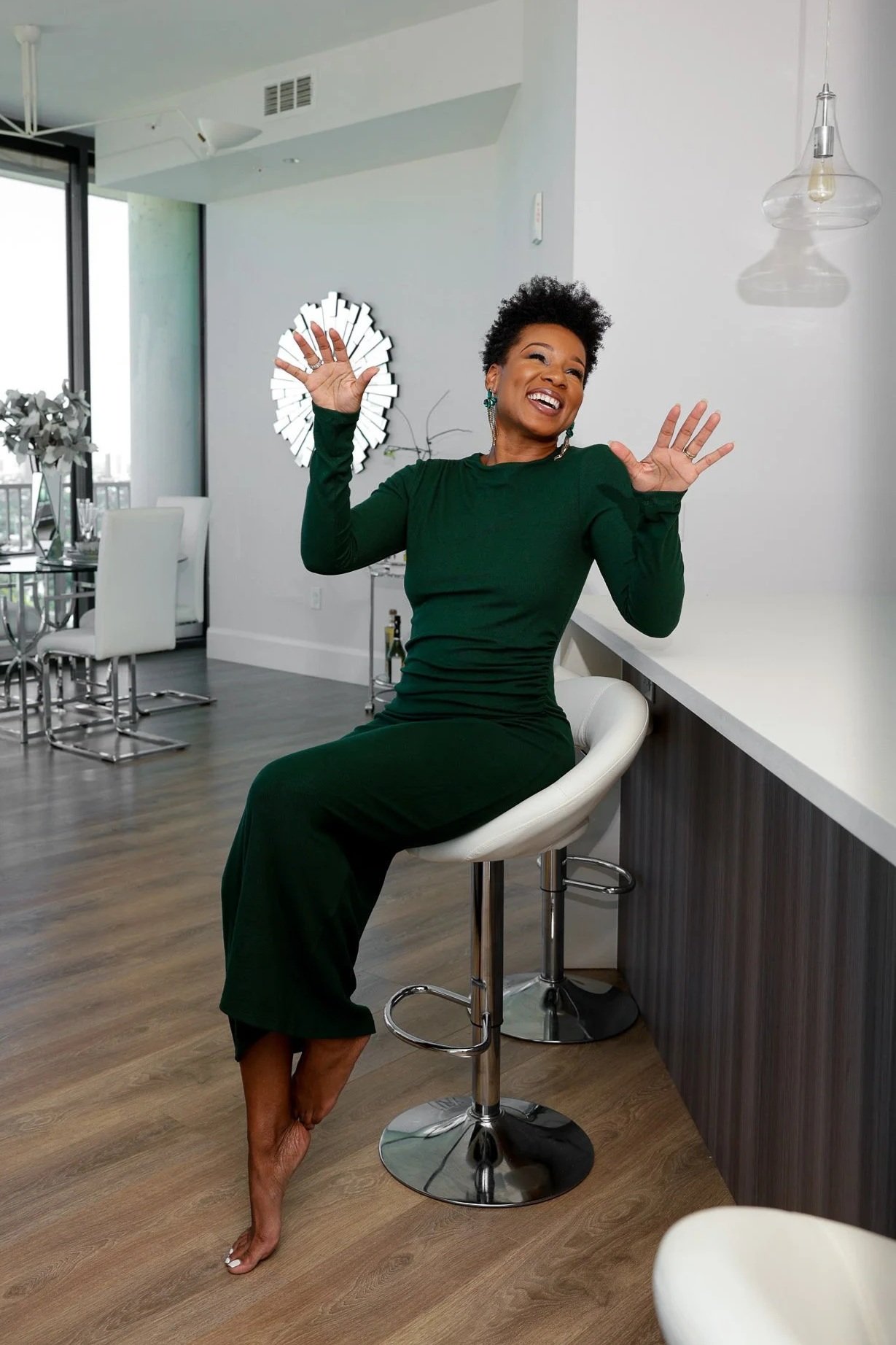 A woman wearing a long emerald-green dress sitting on a bar stool, smiling and gesturing happily with her hands inside a modern, brightly lit apartment.