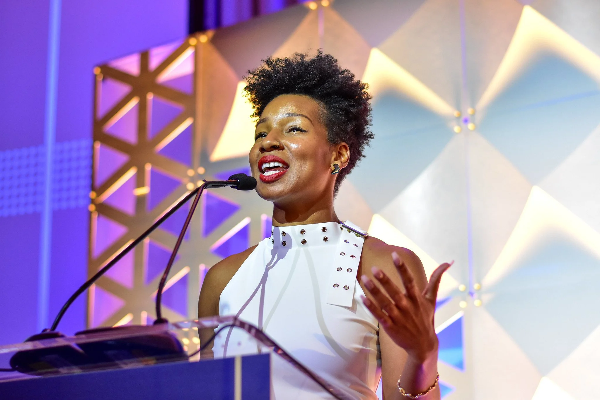 A woman speaking at a podium, smiling, with a microphone, in front of a geometric patterned backdrop with warm-colored lighting.
