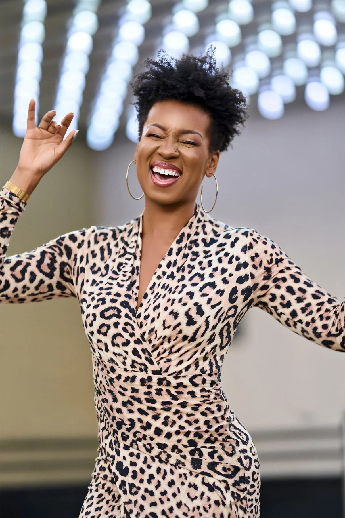 A woman with a big smile, wearing a leopard print dress and hoop earrings, appears to be dancing or celebrating.