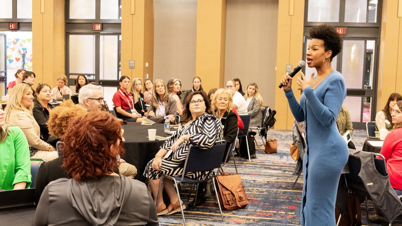 A woman in a blue dress giving a speech or presentation to an audience in a conference room with tables and attendees listening attentively.