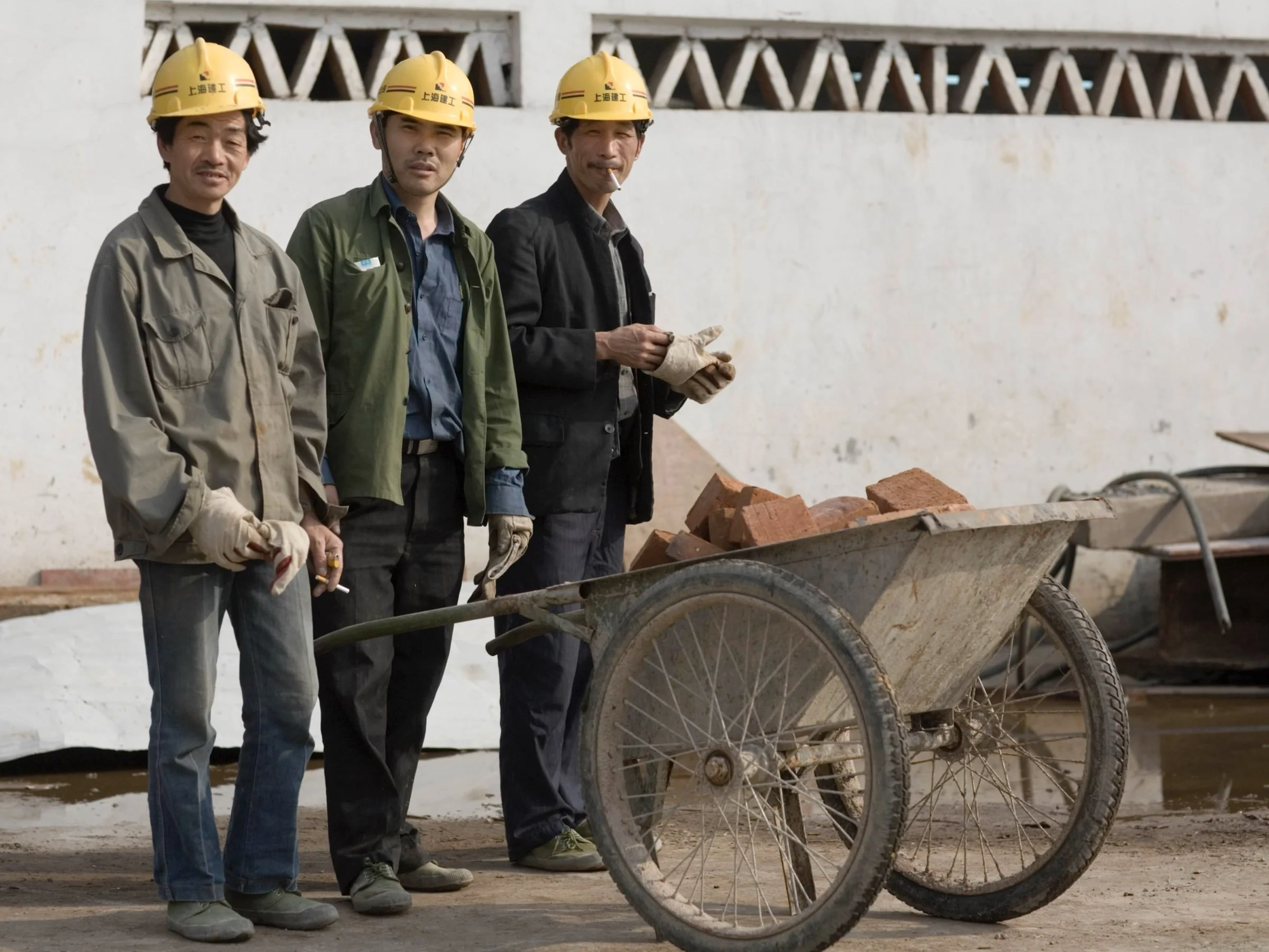 Three construction workers in Henan province posing with a wheelbarrow and bricks wearing yellow hard hats and Ospop workwear.