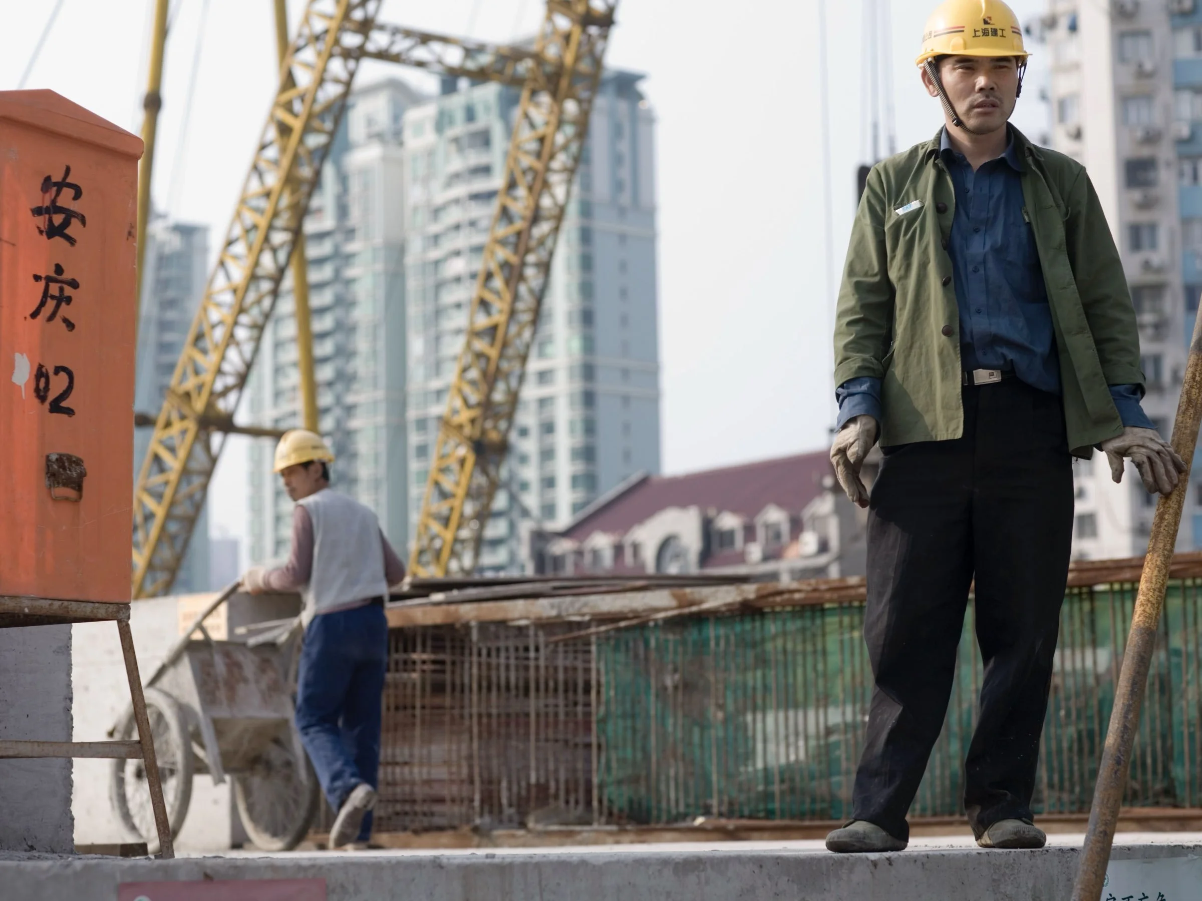 Portrait of a Chinese construction worker standing on a concrete ledge with a crane and high rise apartments in the background.