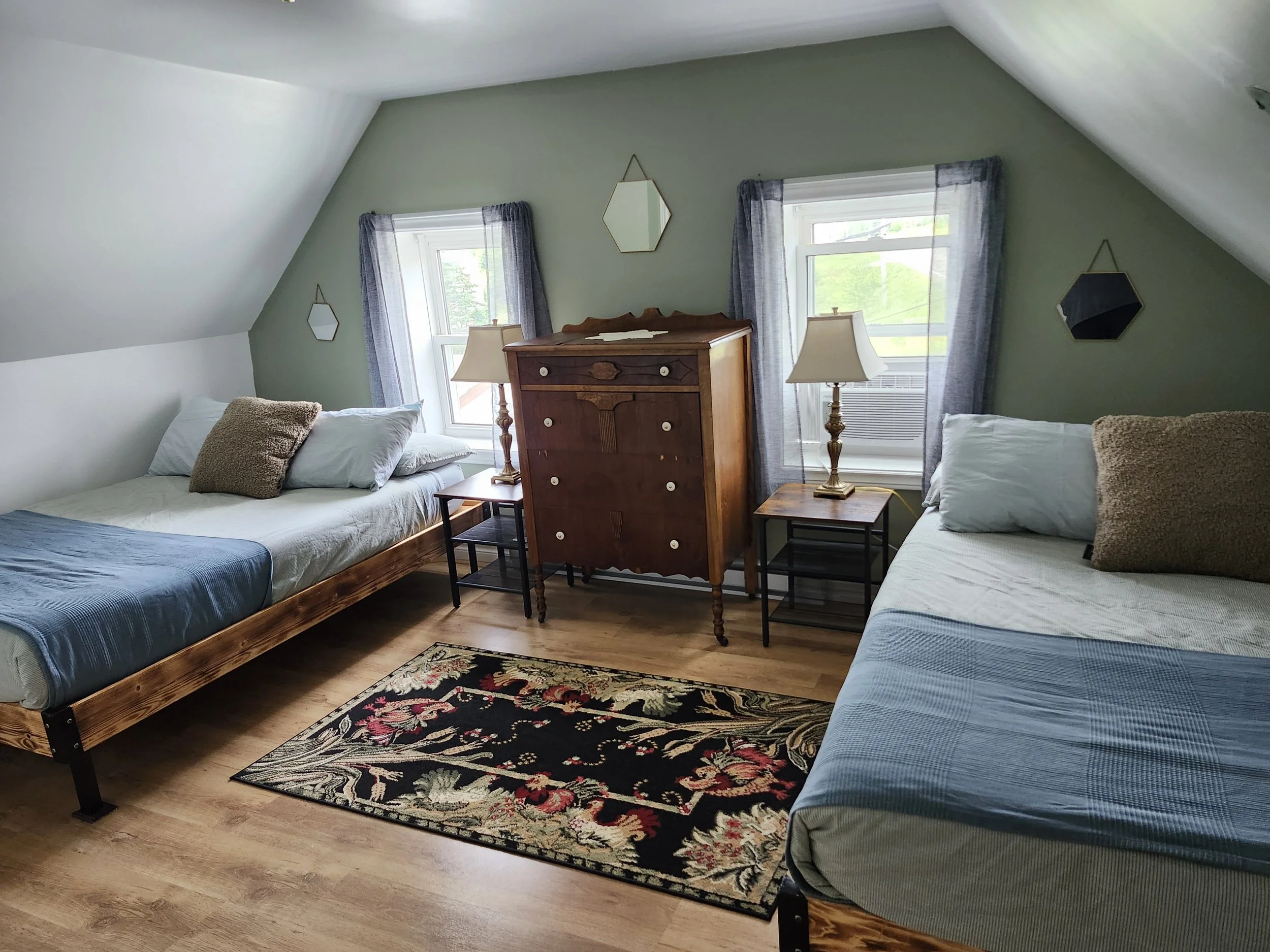 A cozy bedroom with two twin beds, a wooden dresser, two lamps, two windows with blue curtains, and a floral rug on wooden floor.