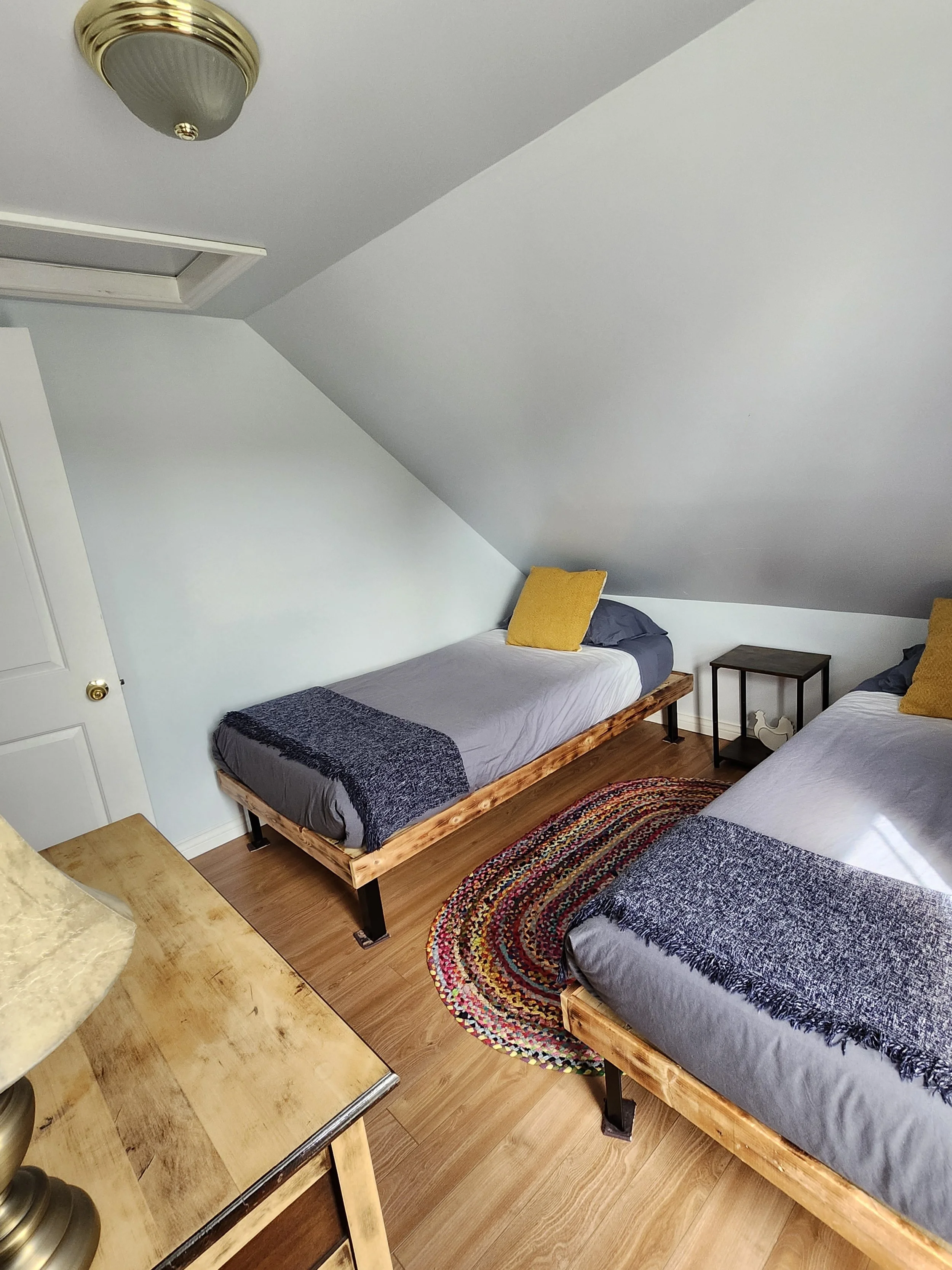 Room with two twin beds with gray bedding and yellow pillows, a black nightstand between them, a colorful braided rug, ceiling light, part of wooden dresser, and sloped ceiling.