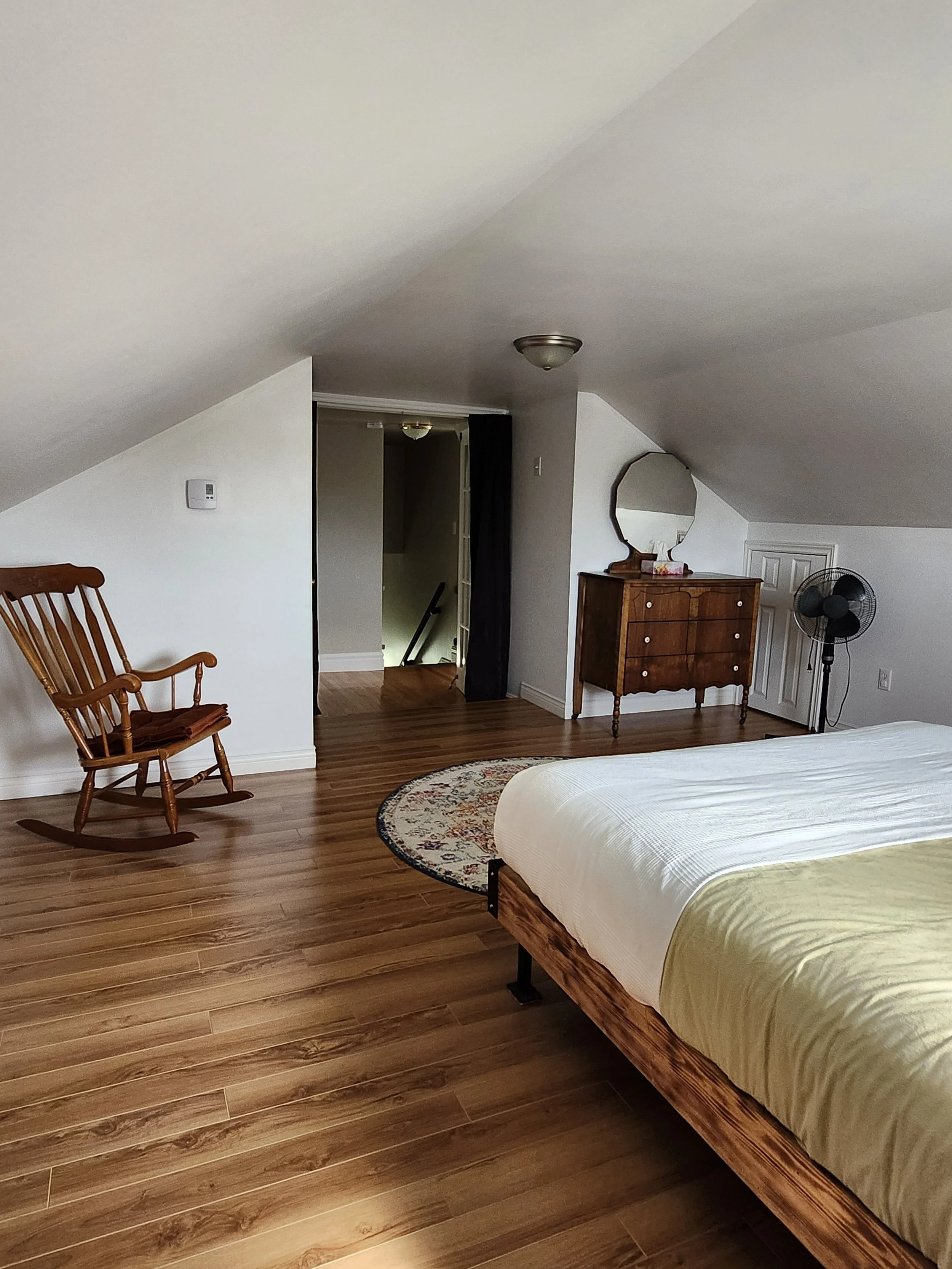 A bedroom with a sloped ceiling, hardwood floors, a bed with a white and yellow bedspread, a wooden rocking chair, a small round rug, a wooden dresser with a mirror, a black fan, and a doorway leading to a staircase.