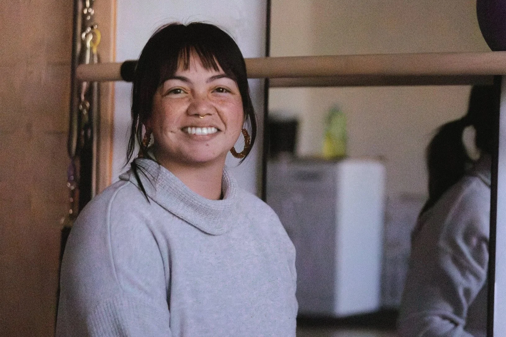 A woman with dark hair, wearing a light gray turtleneck and large hoop earrings, smiling and indoors in a yoga studio.