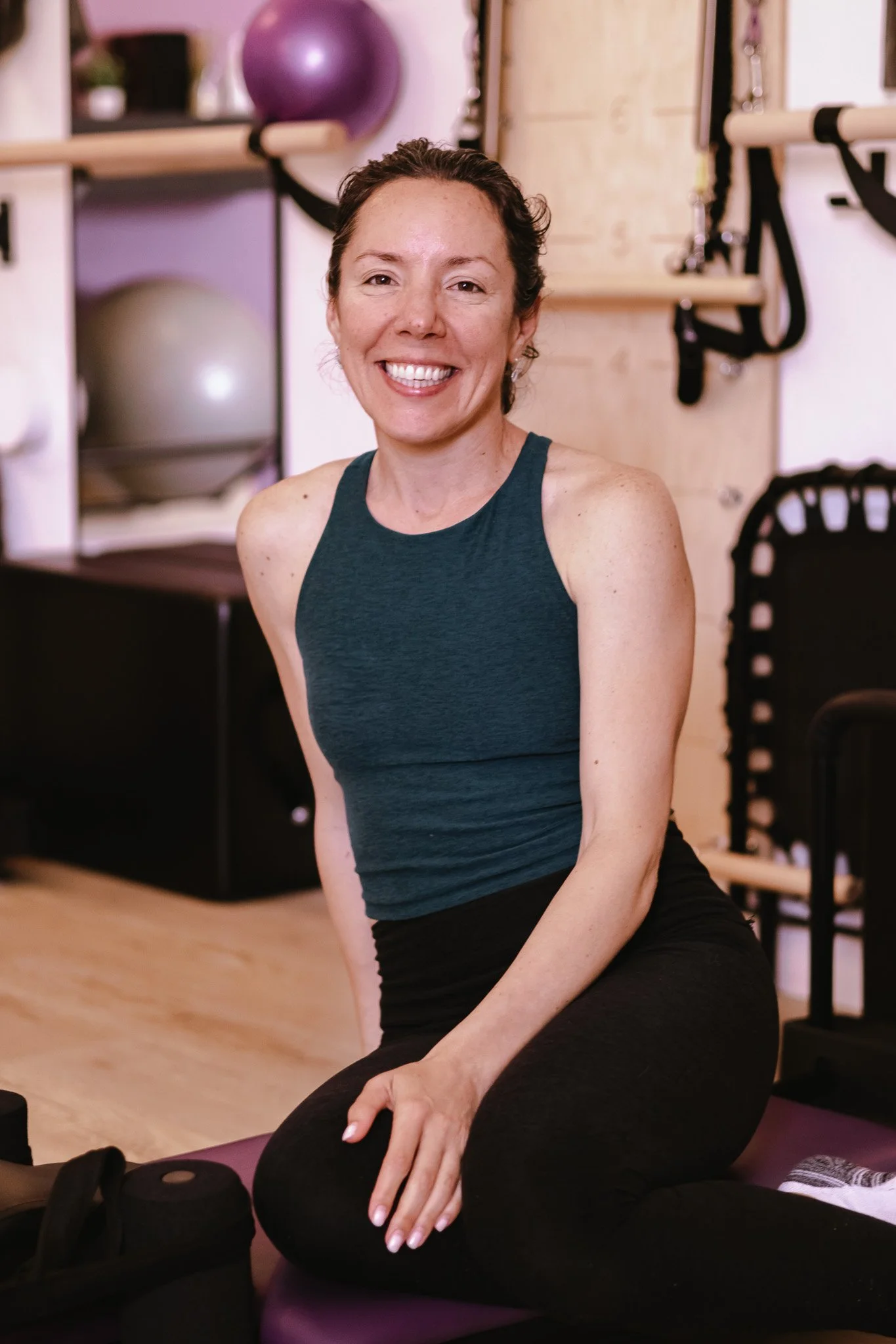 A woman smiling and sitting on a purple exercise mat in a gym or fitness studio, with exercise equipment and stability balls in the background.