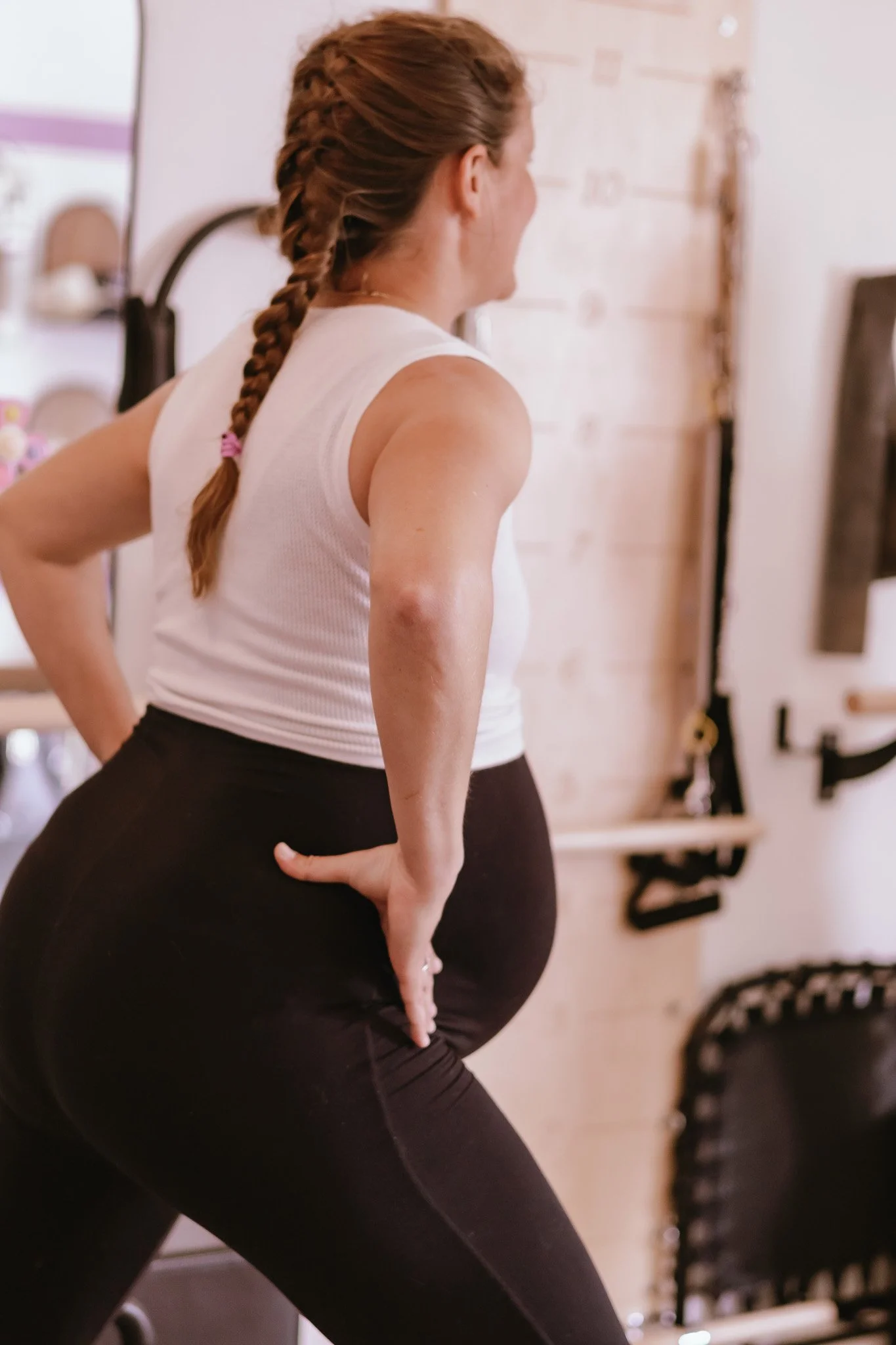 Pregnant woman with braided hair stretching in a fitness studio, touching her lower back with one hand, wearing a white sleeveless top and black leggings.