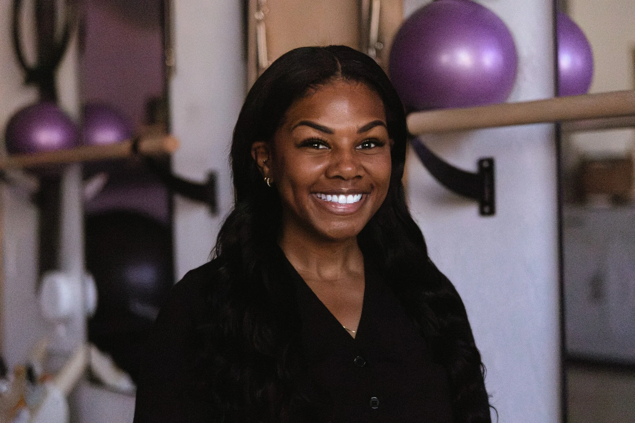 A woman with dark, wavy hair and a bright smile, wearing a black top. She is in a room with purple exercise balls on shelves behind her.