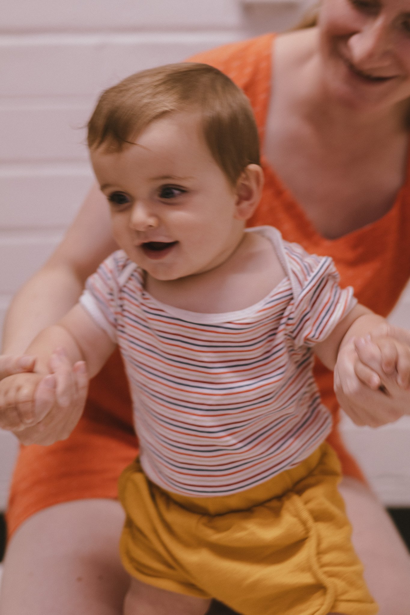 A joyful baby in a striped shirt sitting on an adult's lap, with hands held by the adult, both smiling.