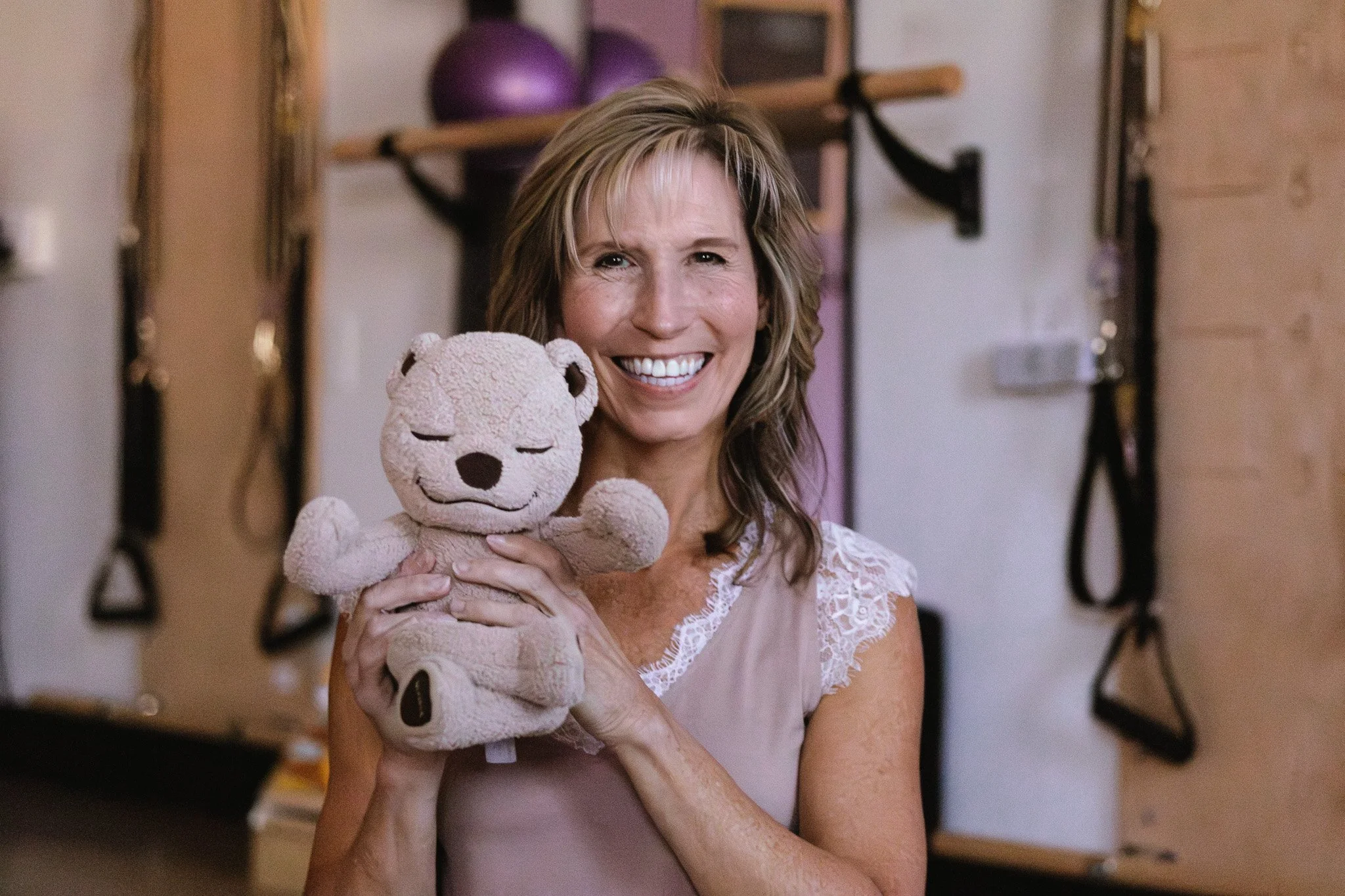 A woman with shoulder-length light brown hair smiling and holding a beige stuffed bear with closed eyes in a kid friendly Pilates & yoga studio with gym equipment in the background.