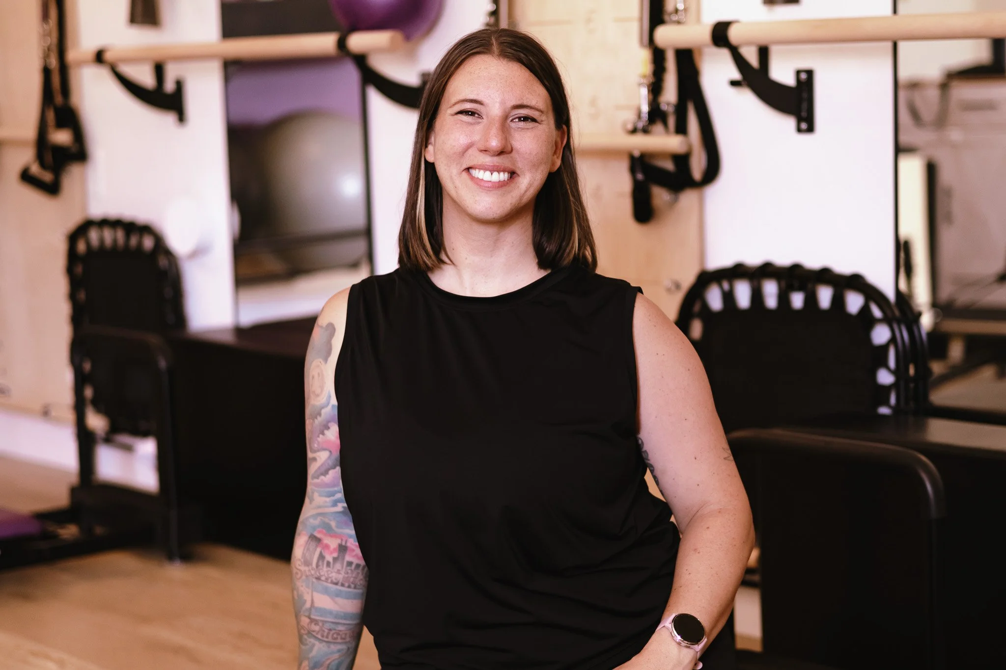 A smiling woman with shoulder-length brown hair, wearing a black sleeveless top and a smartwatch, standing in a gym or fitness studio with workout equipment in the background.