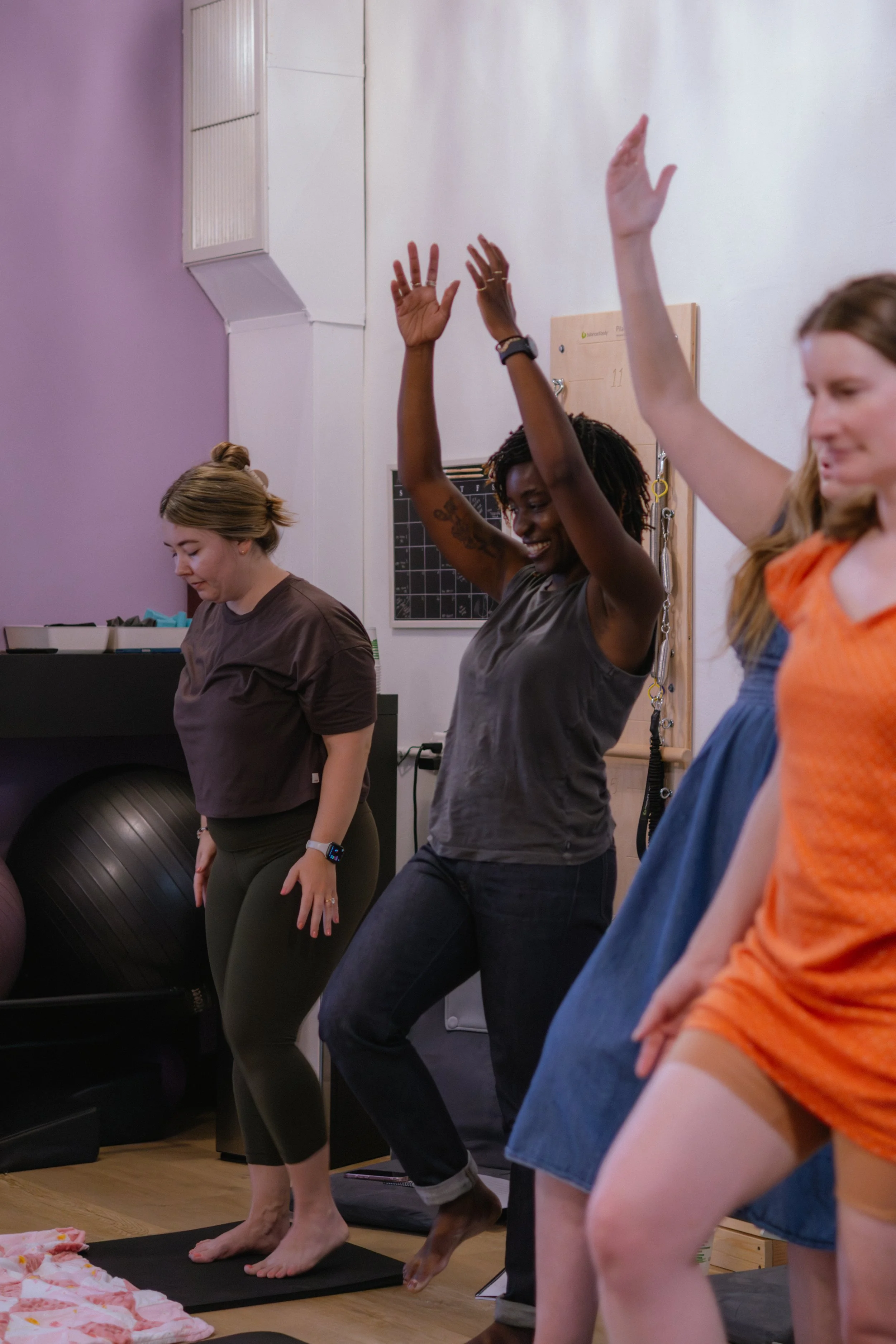 Postpartum women participating in a fitness or yoga class, standing on mats with their arms raised, inside a studio with exercise equipment and a purple wall.