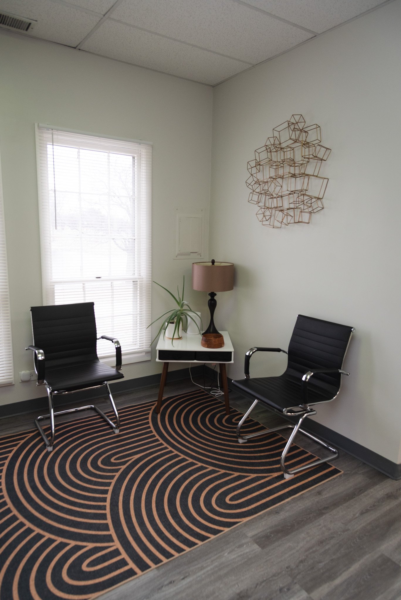Corner of an office with two black chairs, a small white side table with a lamp and plant, a geometric wall art piece, a window with blinds, and a patterned rug on a wooden floor.