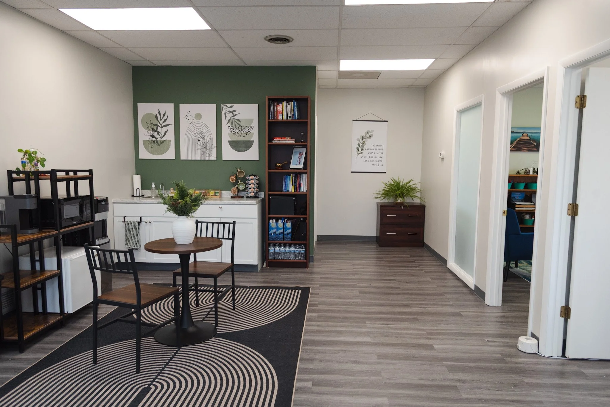 Interior of an office or counseling room with a small round table, three chairs, bookshelves, wall art, plants, and a patterned rug.