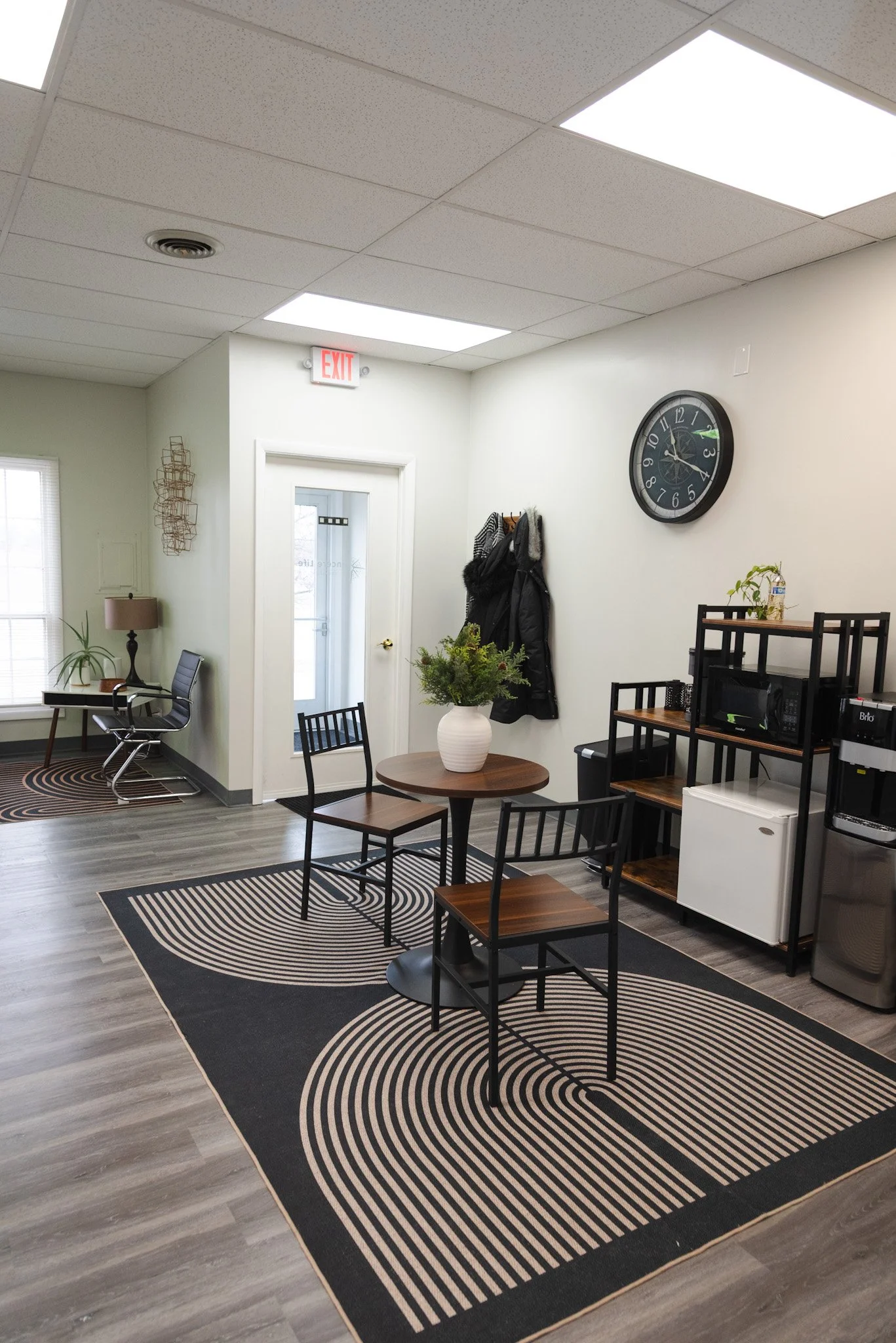 An office break room with a small round table with three chairs, a rug with geometric patterns, a kitchenette with microwave, mini fridge, and shelves, and a coat rack with jackets. There is a clock on the wall and a window near a desk with a chair.