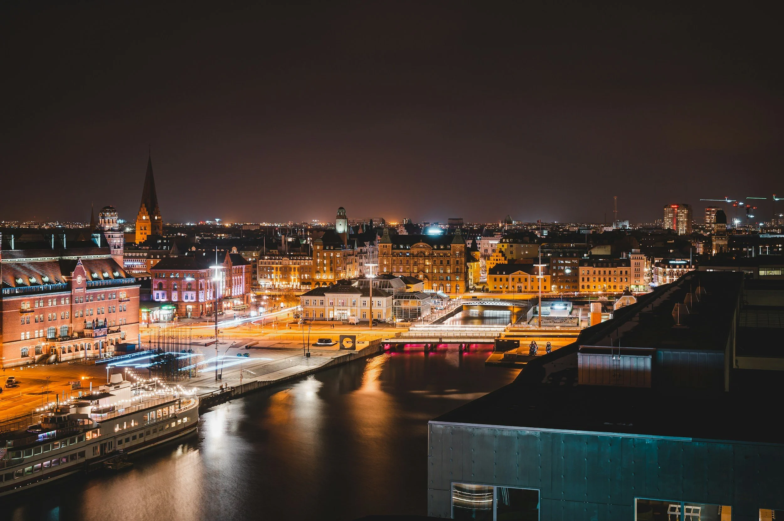 Nighttime cityscape of a European city with illuminated historic buildings, a river, and a bridge.