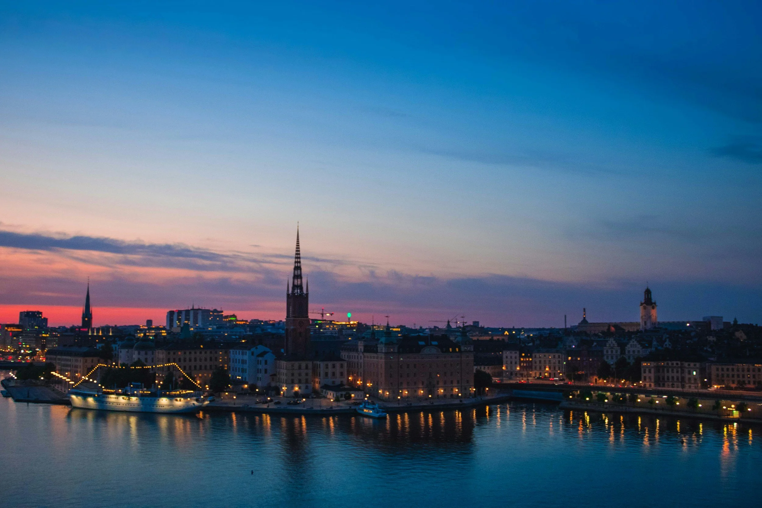 Sunset view of a city skyline along a river with church steeples and historic buildings, illuminated by city lights.