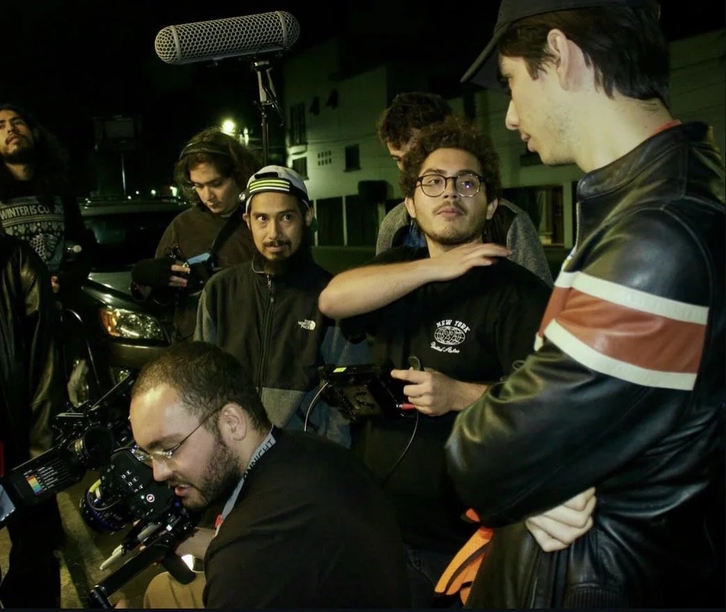 A group of young men, some with cameras and equipment, gathered outdoors at night, with one person appearing to give instructions or discuss a shot.