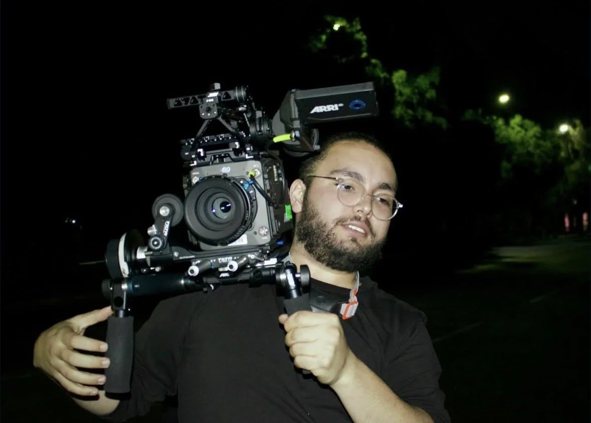 A man with glasses and a beard carrying a professional film camera on his shoulder during nighttime, with dark trees and lights in the background.