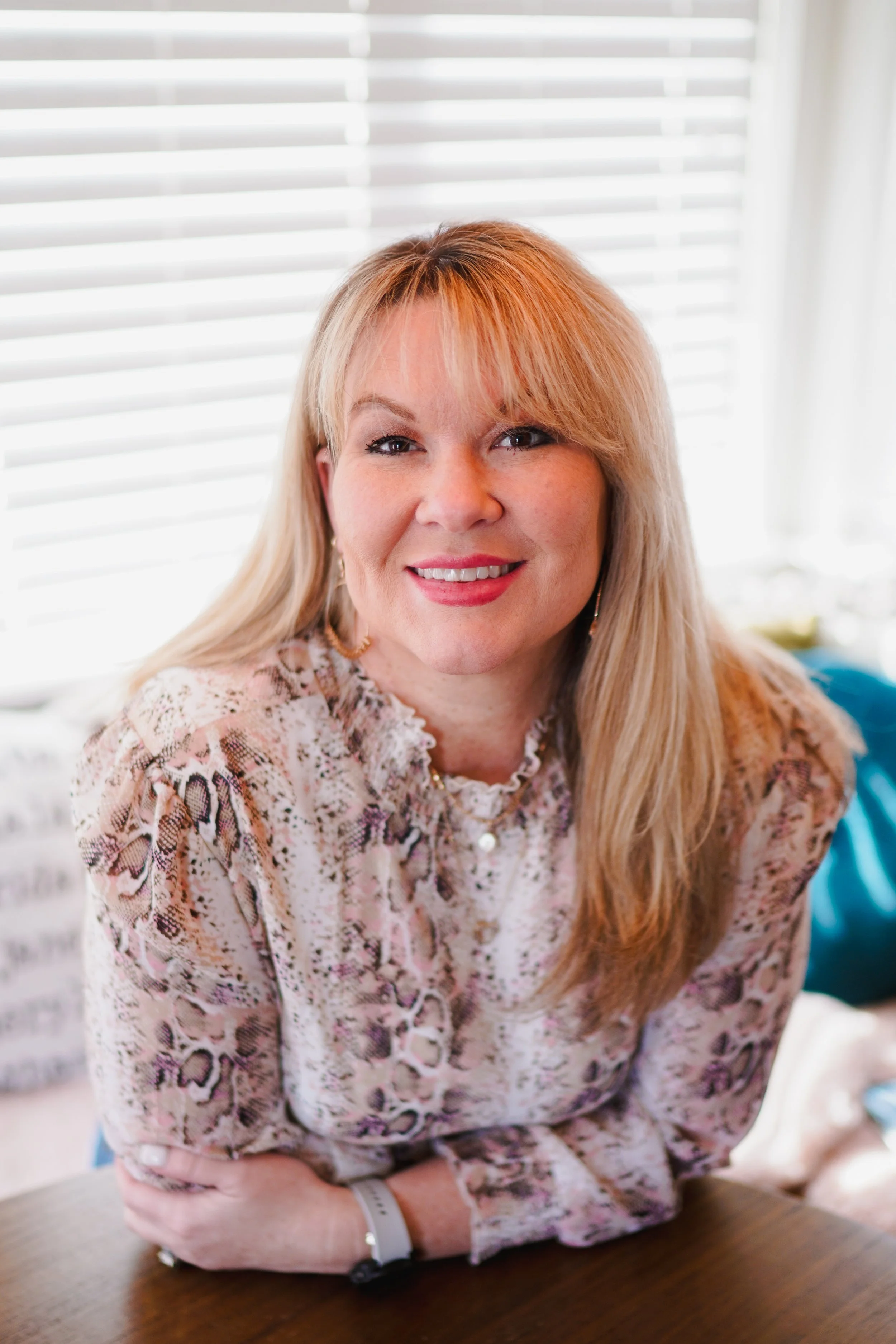 A woman with long blonde hair and a pink patterned blouse sitting at a table in front of a window with blinds, smiling at the camera.