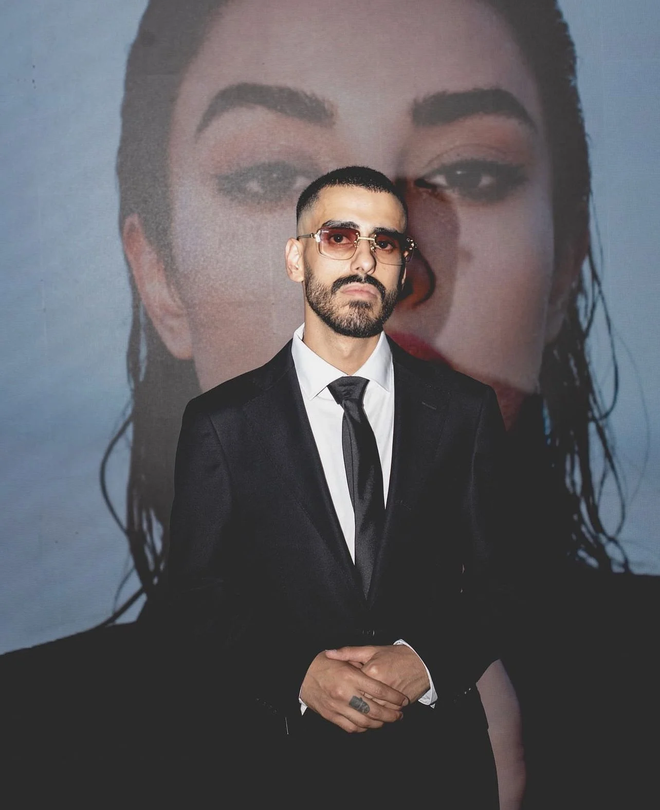 A man in a black suit, white shirt, and black tie standing in front of a large portrait of a woman with dark hair and makeup.