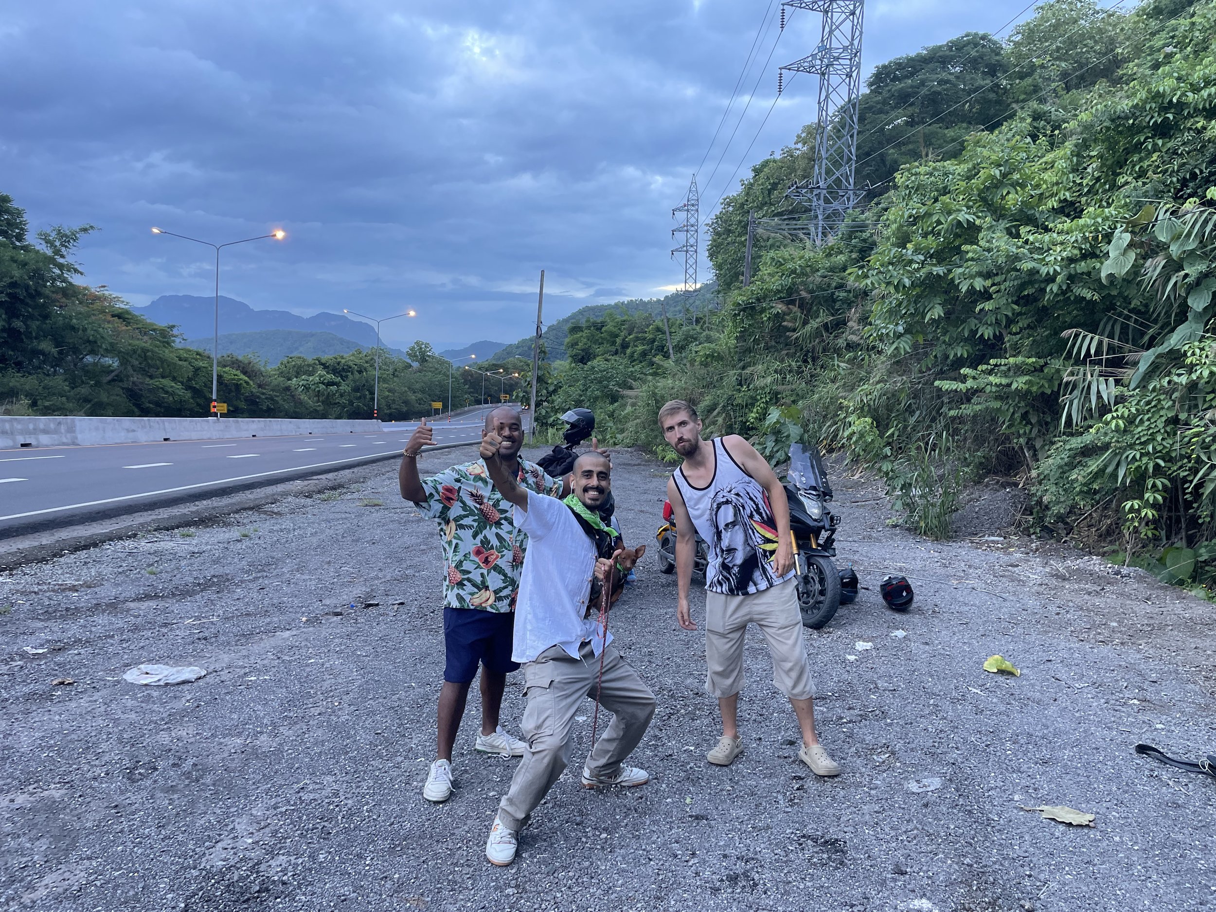Three men standing on a gravel roadside next to a motorcycle, with lush green trees, mountains in the background, and a cloudy sky overhead.