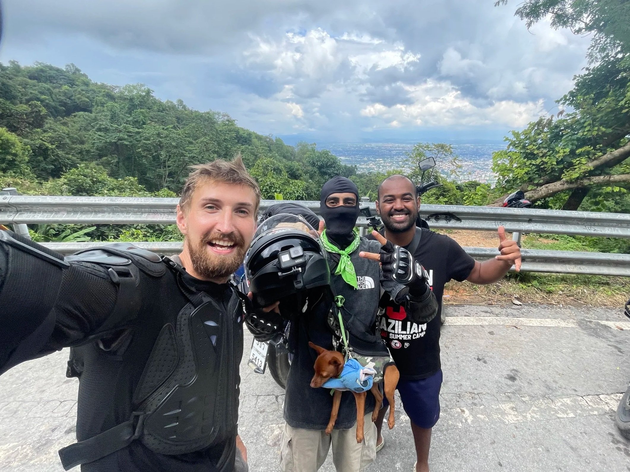 Three smiling men posing for a photo on a mountain road with greenery and a city view in the background, two of them making hand gestures, one holding a helmet and a small dog wearing a blue shirt.