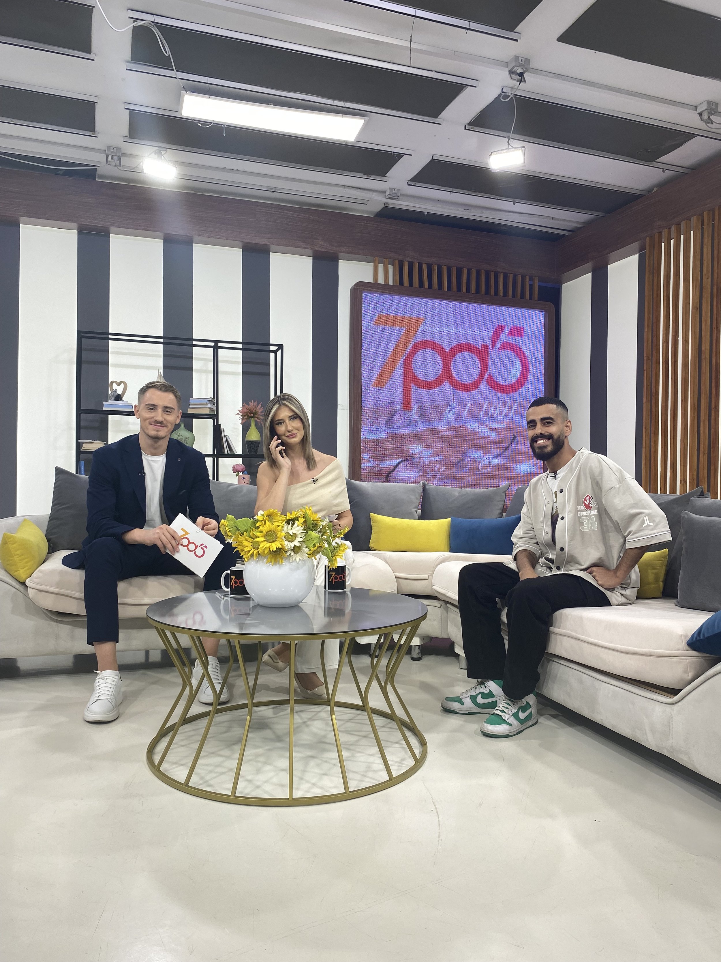Three people sitting on a white sofa on a TV set, with a large screen behind them displaying the logo '7po5'. There is a round glass table with a flower vase and two coffee mugs in front of them. The studio has a modern design with black, white, and wooden accents.