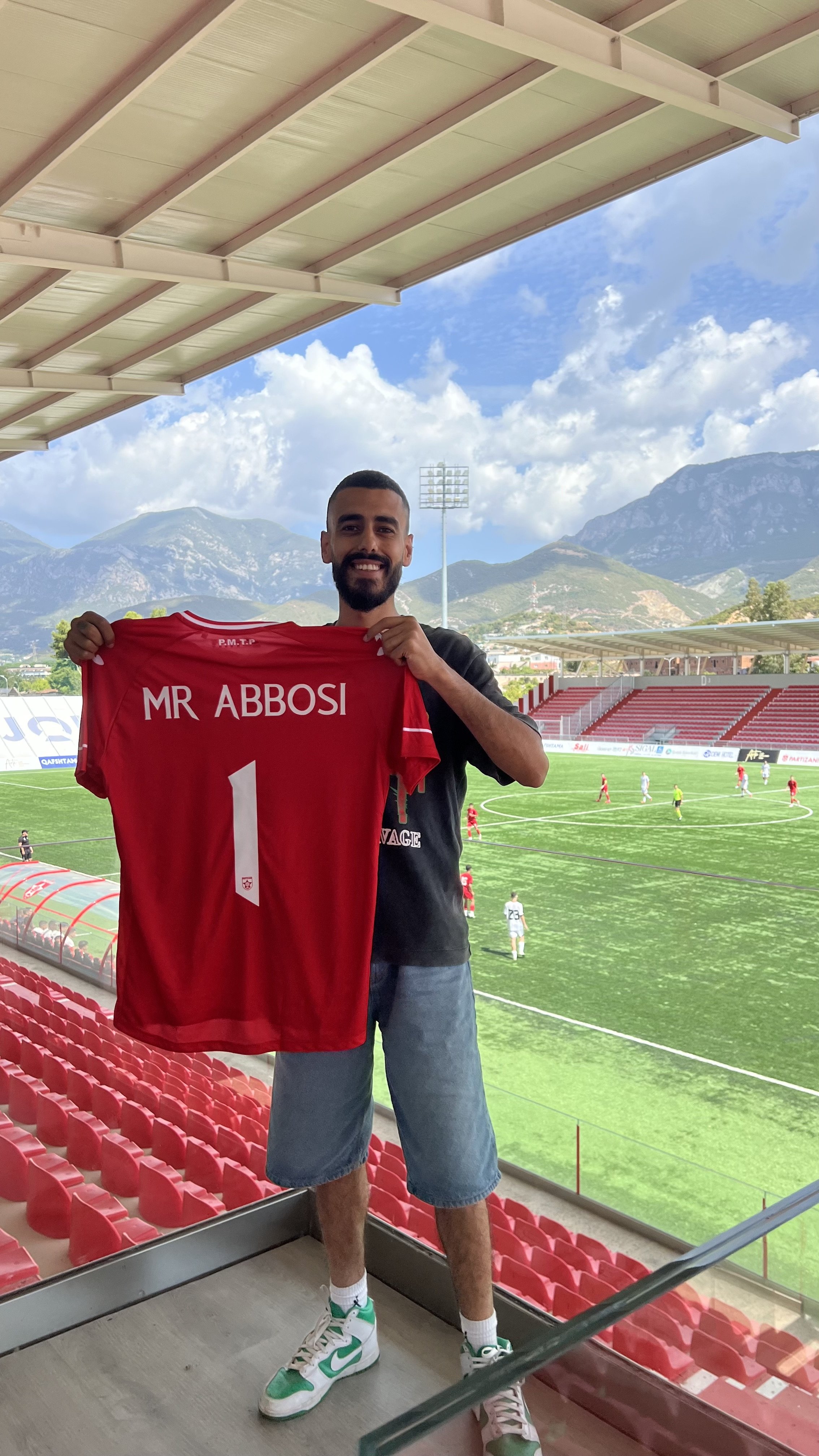 A man with a beard and short dark hair smiling and holding a red soccer jersey with the name 'MR ABBOSI' and the number 1 on it, standing in a soccer stadium with a green field, other players, and a mountainous landscape in the background.