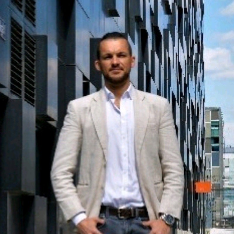 A man standing outdoors in front of a modern building with metal panels and windows, wearing a white blazer, white shirt, and gray pants.