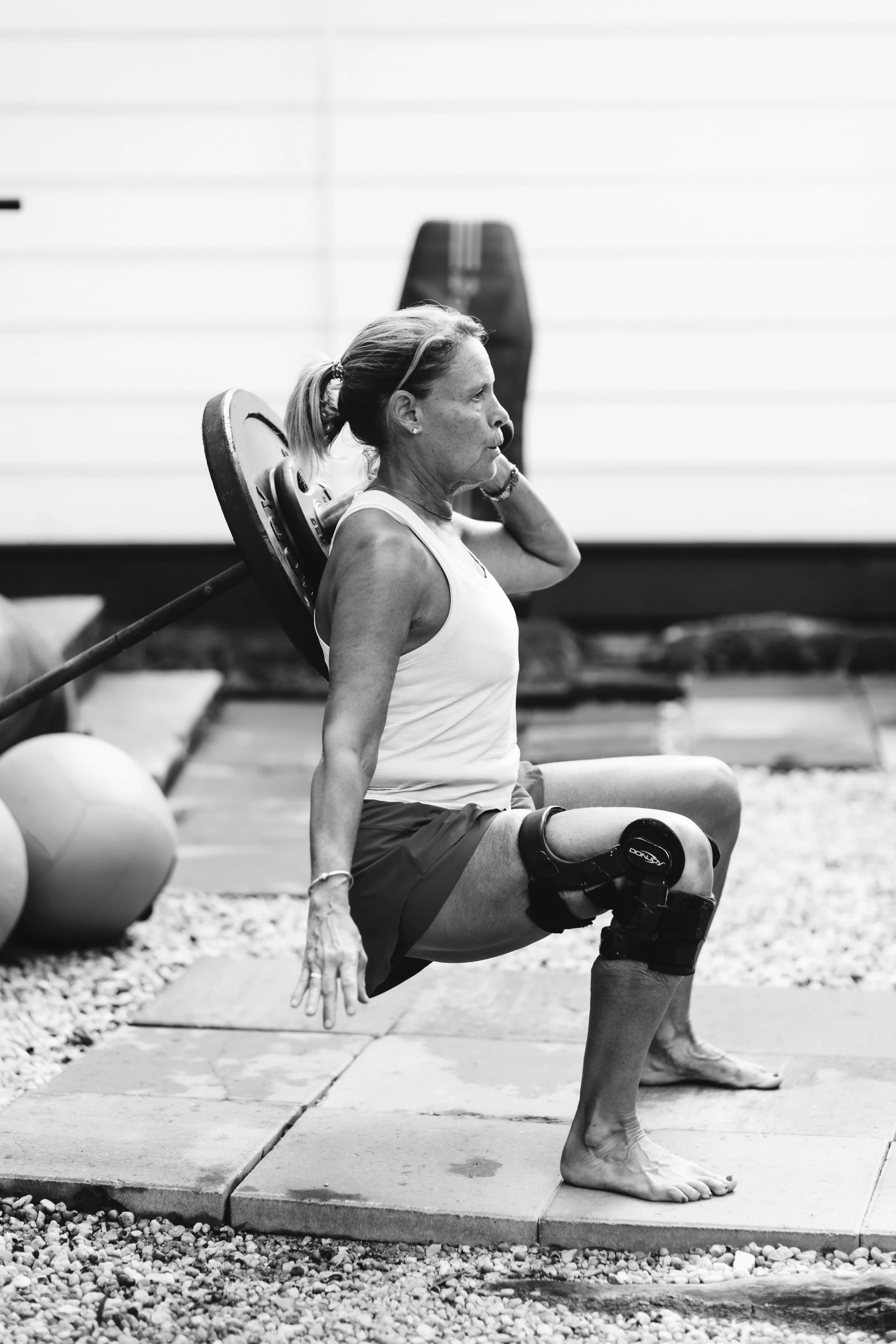 An elderly woman with a prosthetic knee performs a squat exercise outdoors, with a barbell on her shoulders.
