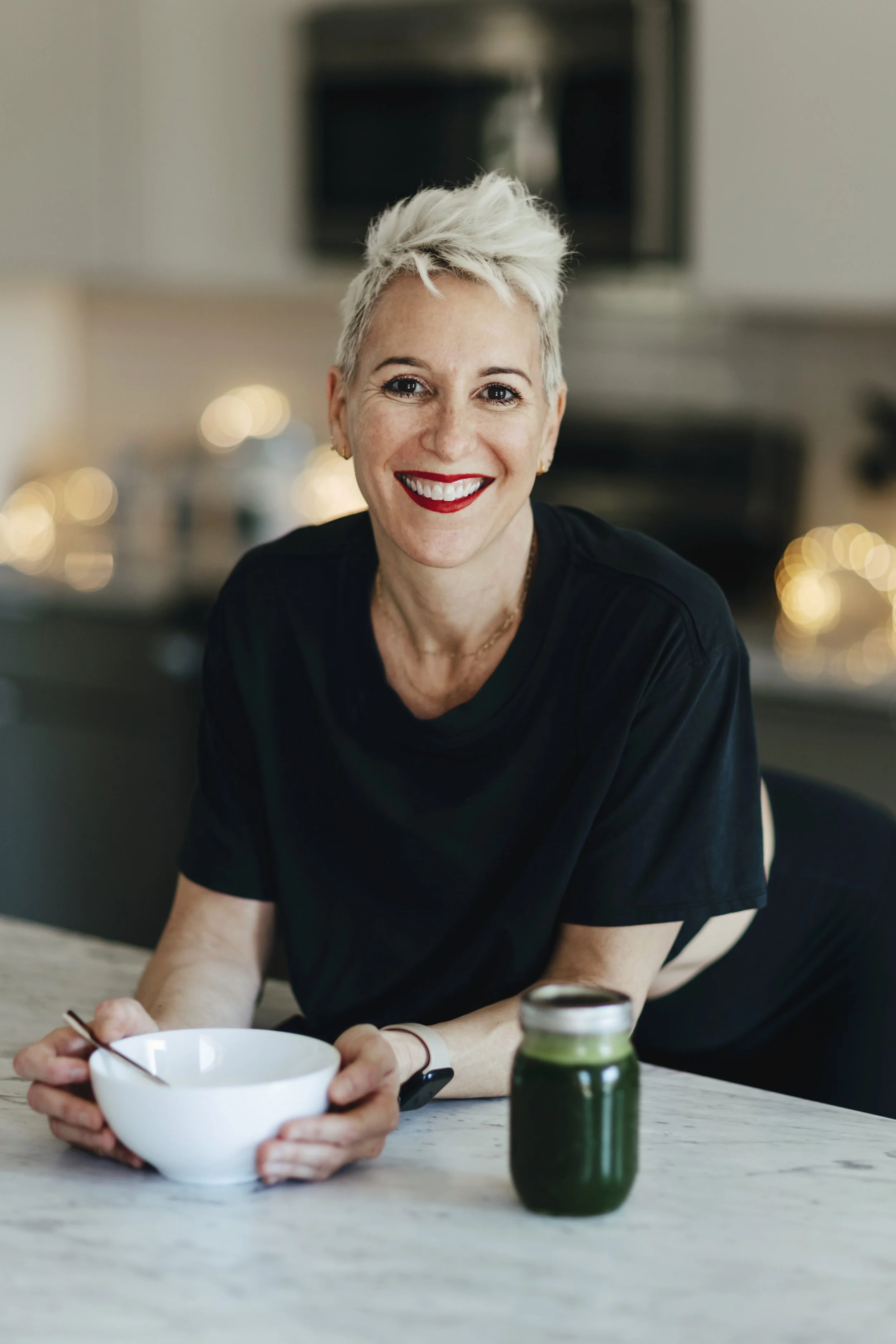 A woman with short, platinum blonde hair and red lipstick smiling at the camera while holding a white bowl and spoon, sitting at a table with a jar of green juice in front of her.