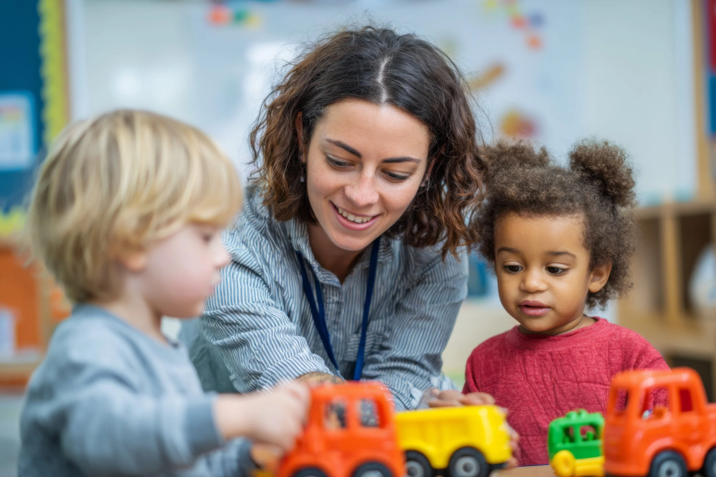 A young female teacher engaging with two children, a blonde boy and a girl with curly hair, playing with colorful toy trucks in a classroom.