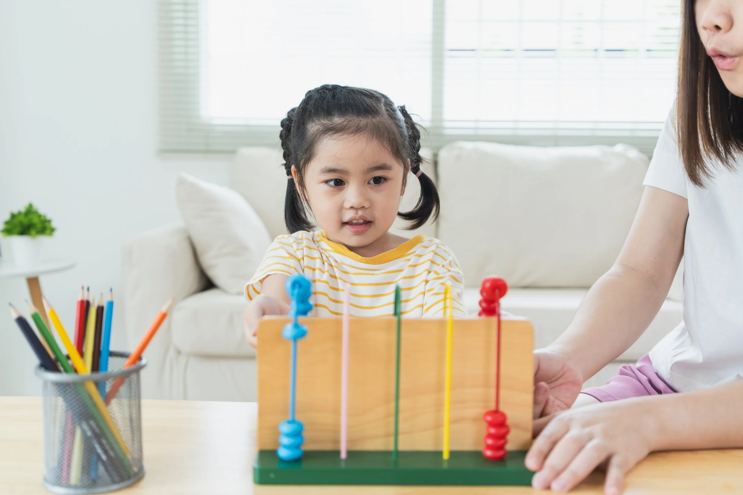 A young girl with dark hair in pigtails and wearing a yellow and white striped shirt playing with an abacus while an adult assists her at a table, in a bright living room.
