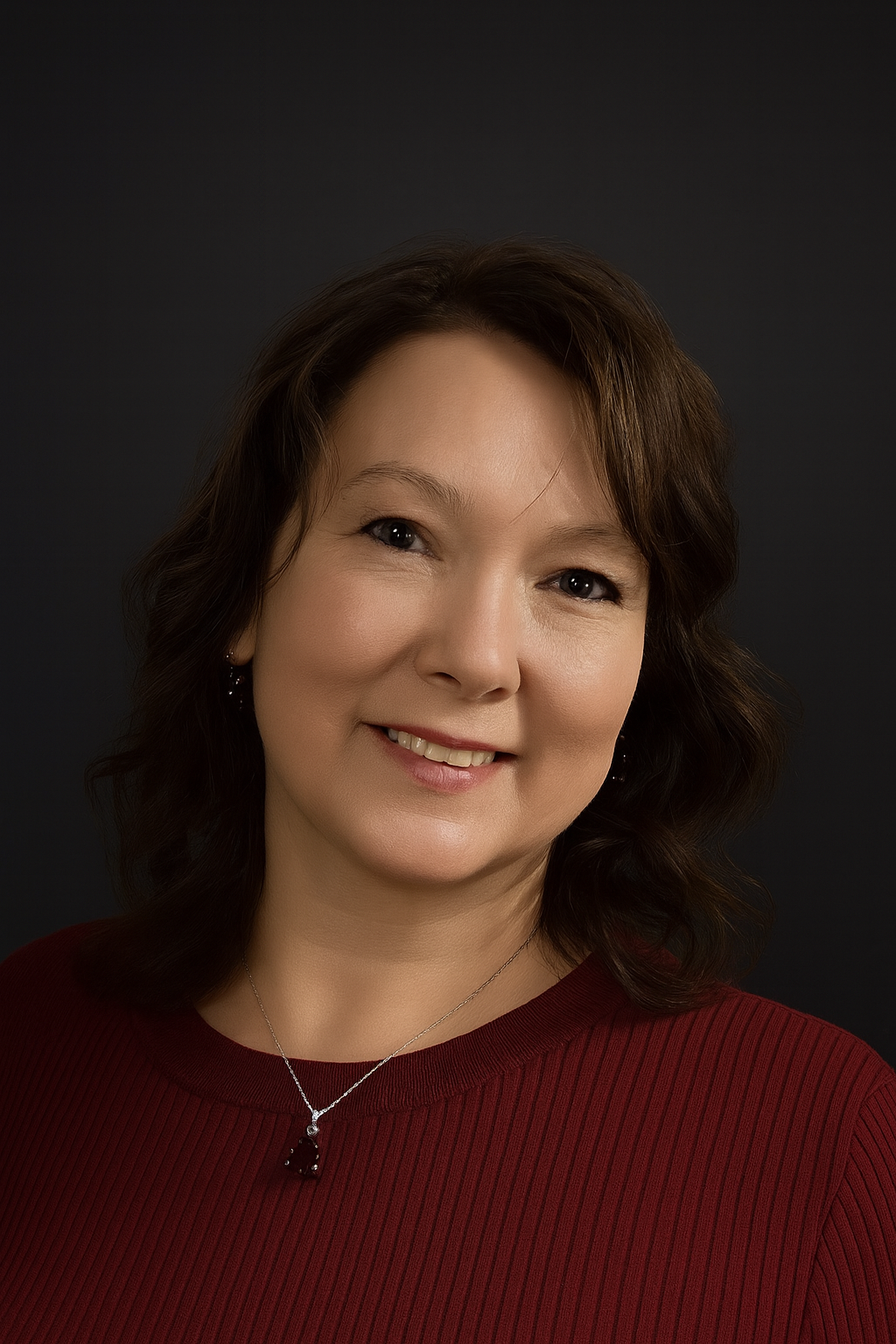 A woman with shoulder-length brown hair wearing a red top and a silver necklace with a dark pendant, smiling against a dark background.