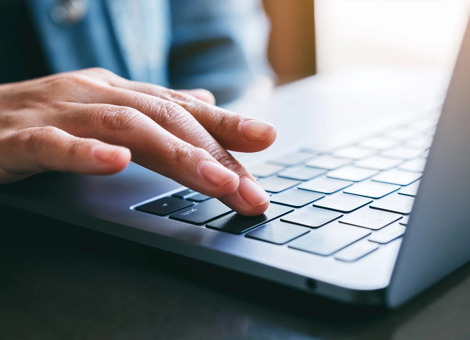 Close-up of a person's hand typing on a laptop keyboard with sunlight coming from the right.