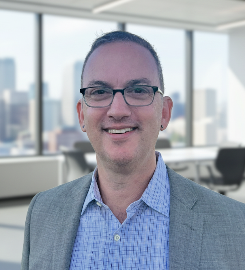 A man wearing glasses and a grey suit smiling in a modern office with large windows and a city skyline in the background.