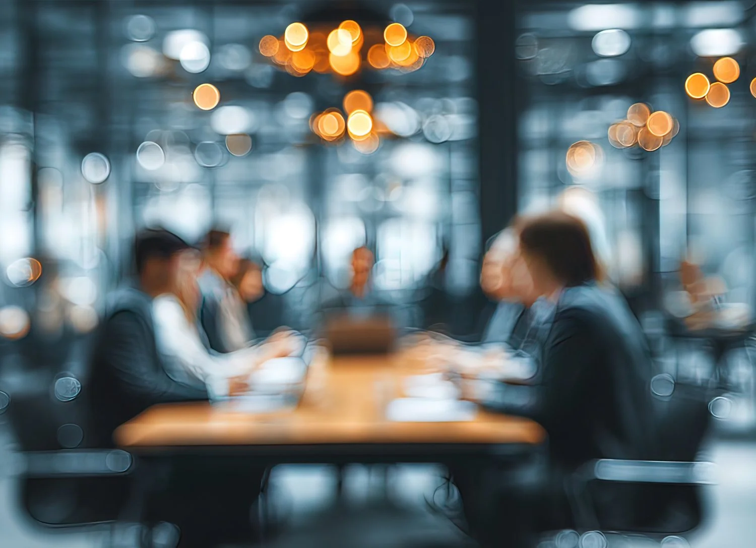 Blurred image of a business meeting in a modern office, with four people sitting at a table, talking and working on laptops.