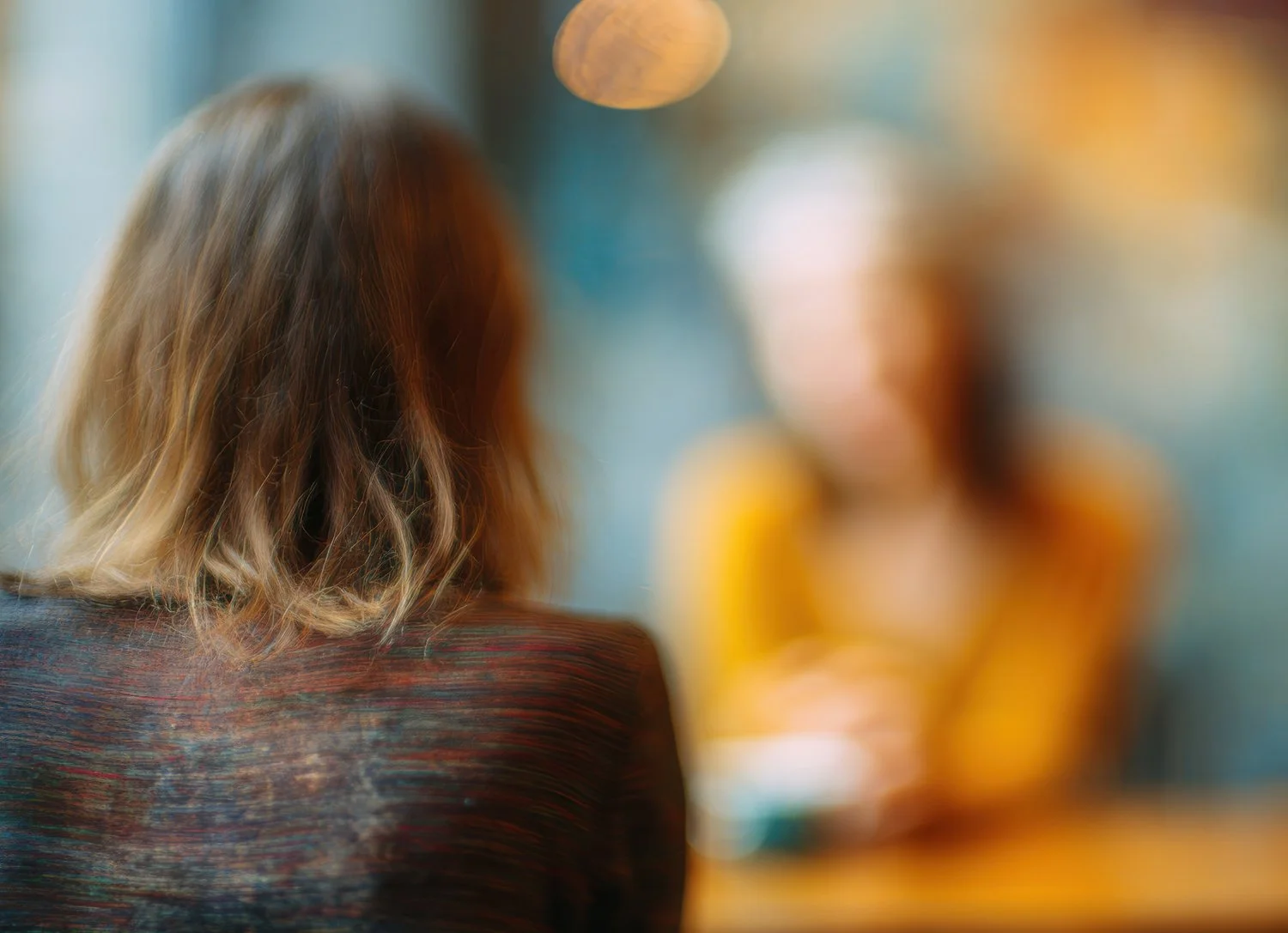 Blurred image of women sitting in a casual conversation or meeting, with focus on woman with wavy blond hair seen from the back.