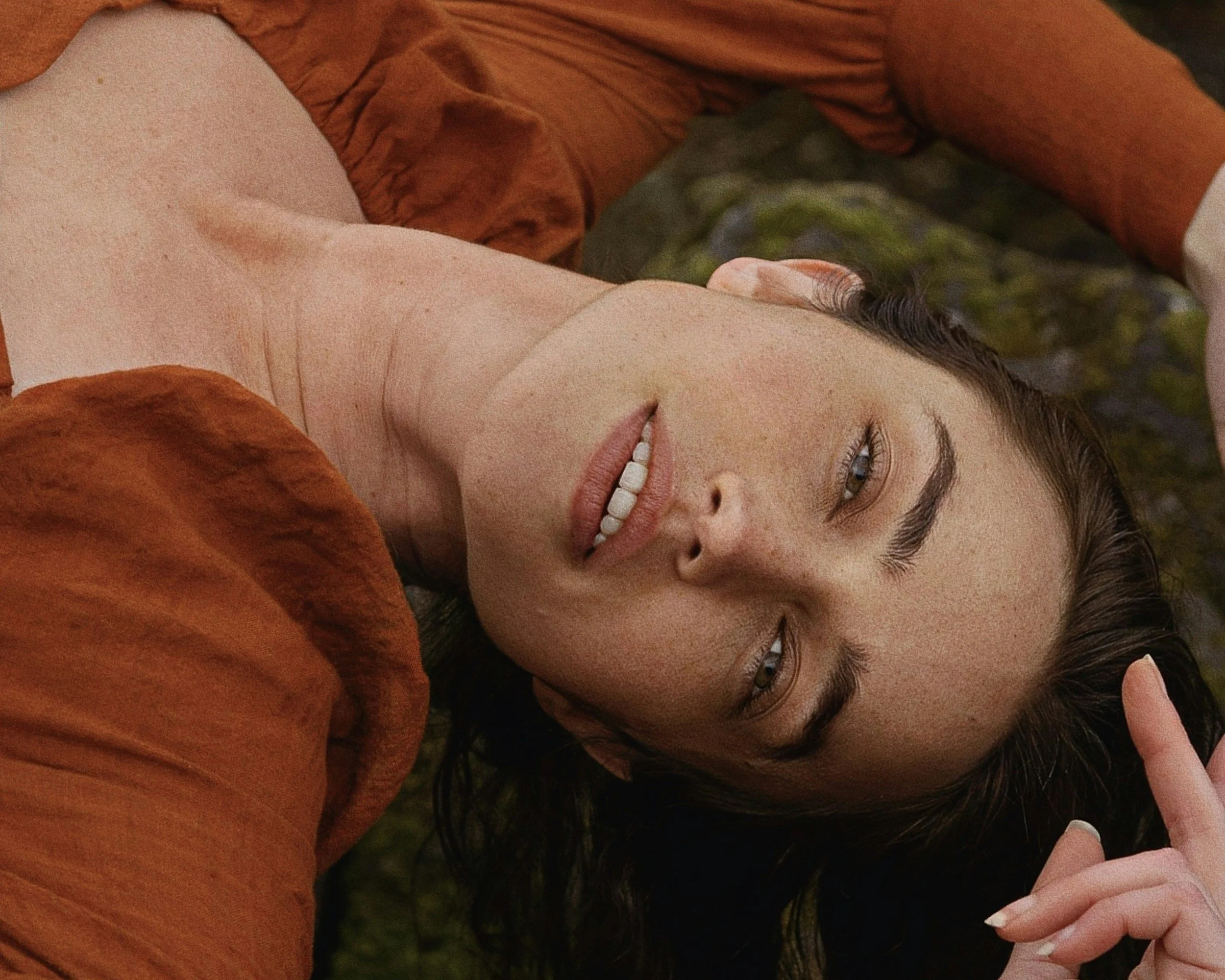 A woman with long dark hair, lying on the ground, wearing a rust-colored jacket, smiling softly at the camera with a hand touching her forehead.