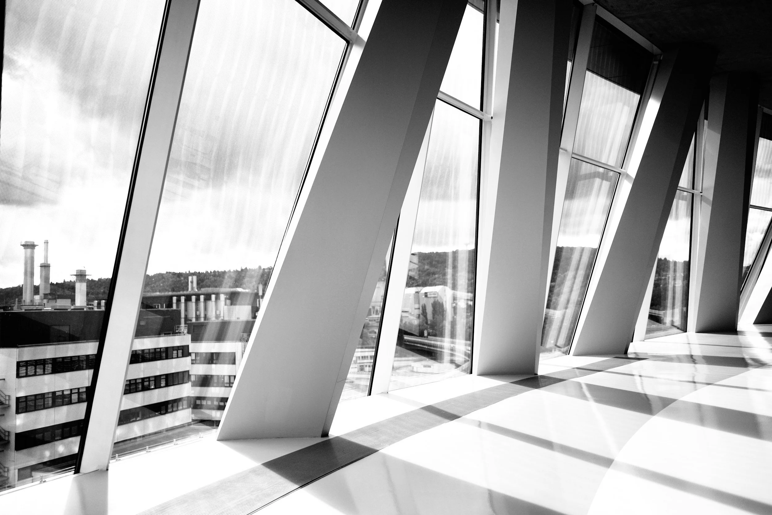 Modern interior with large, angled floor-to-ceiling windows allowing natural sunlight to cast shadows on the floor, overlooking an urban cityscape.