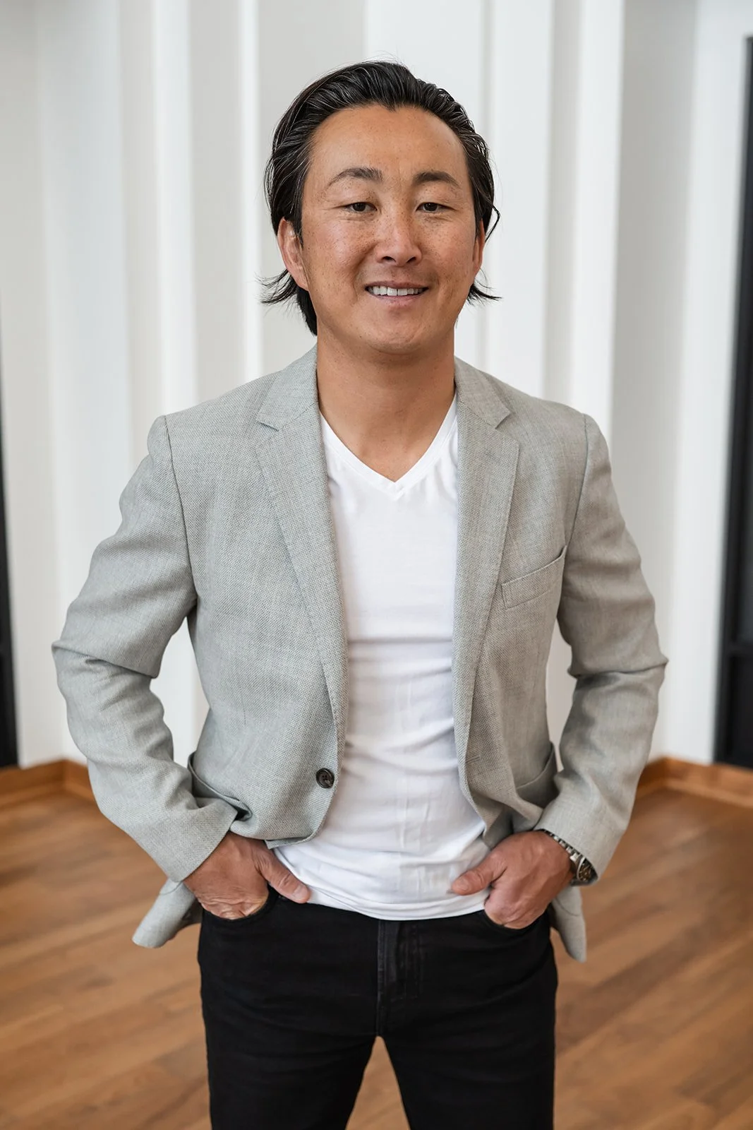 A man with dark, slicked-back hair wearing a light gray blazer, white t-shirt, and black pants, standing indoors with hands in pockets and smiling.