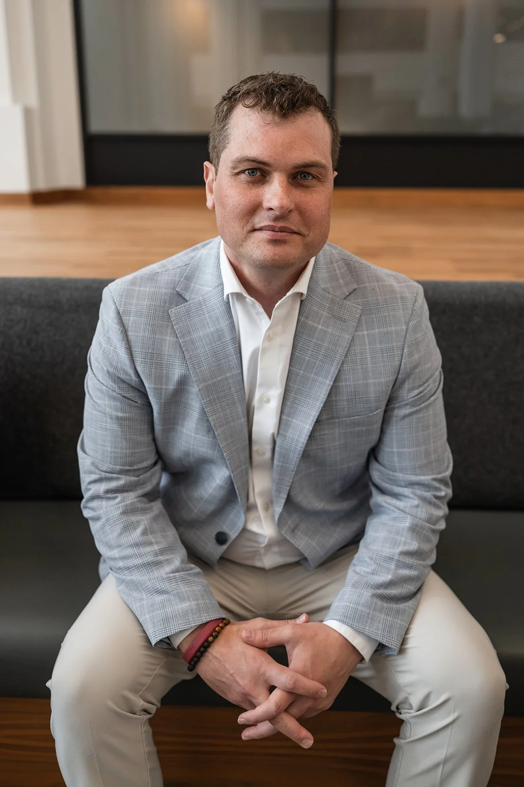 A man with short brown hair and blue eyes sitting on a dark gray bench, wearing a light gray plaid blazer, white dress shirt, and beige pants, in an indoor setting with wooden flooring and modern decor.