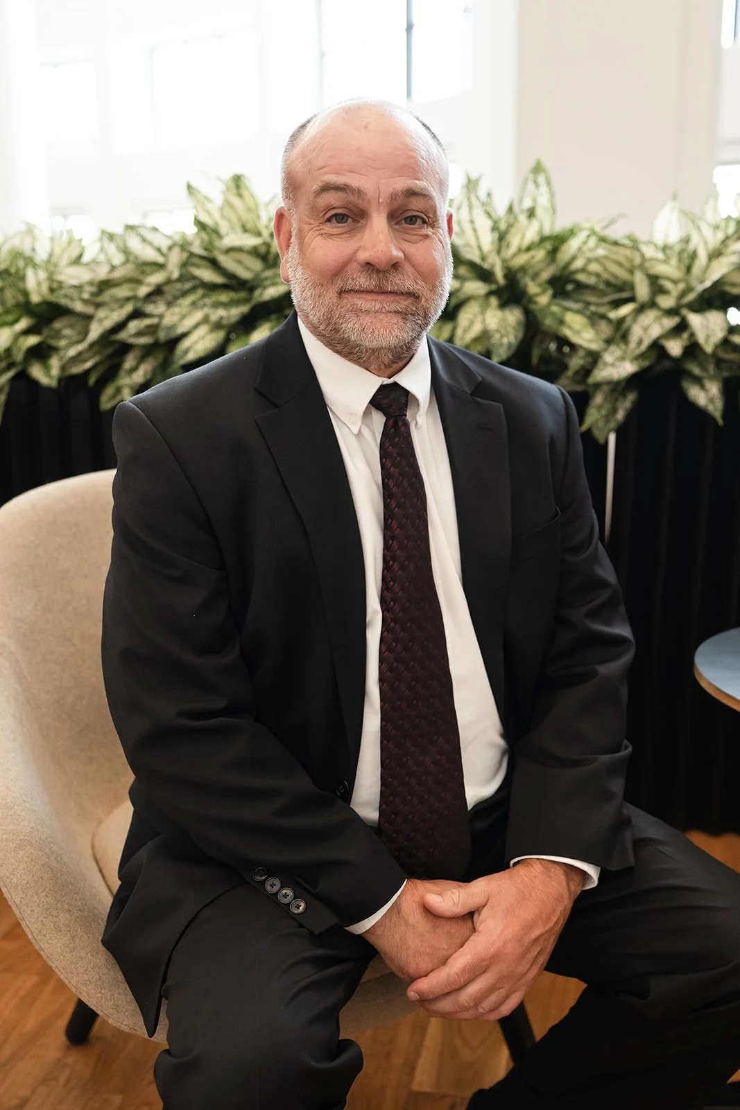 A middle-aged man with a beard and bald head, wearing a black suit, white shirt, and maroon tie, sitting on a beige chair with greenery in the background.