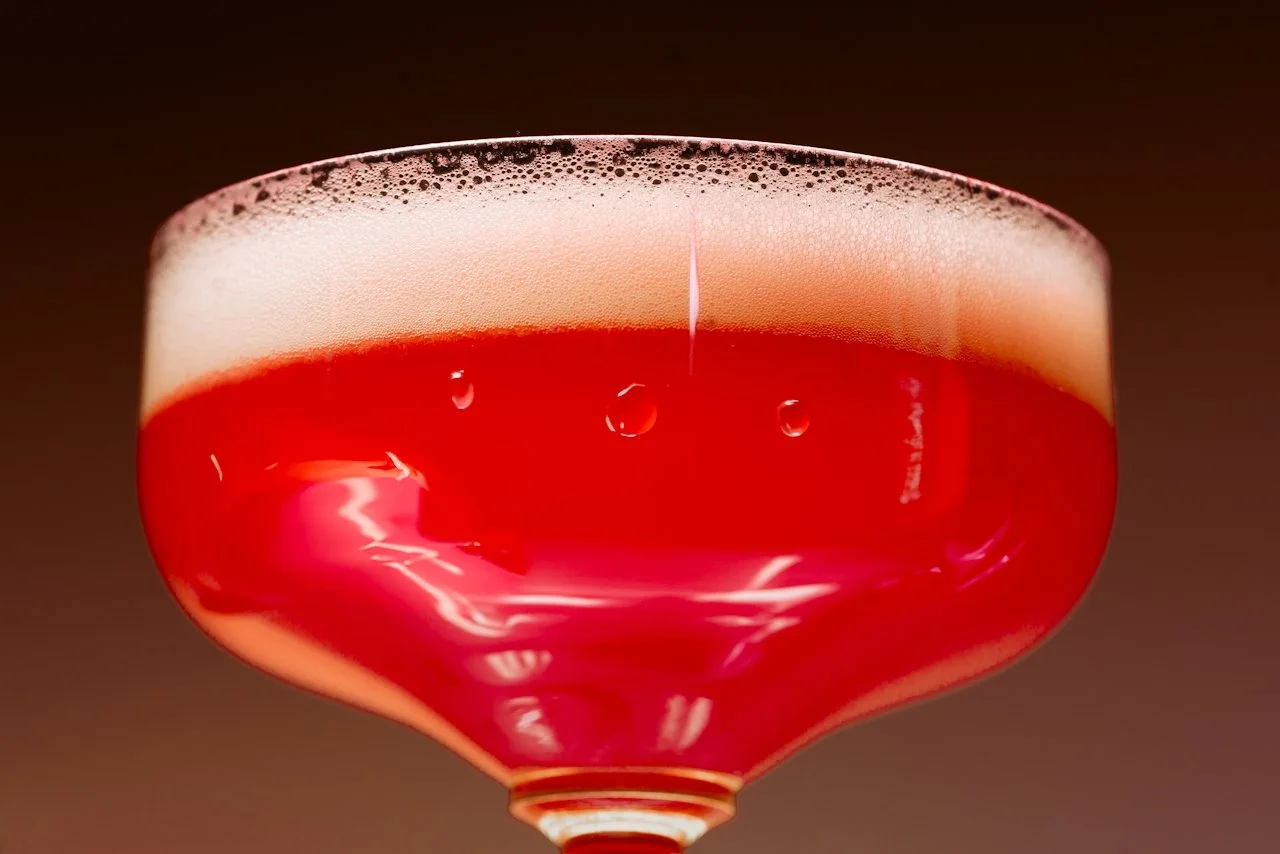 Close-up of a red cocktail in a pink coupe glass with foam on top and bubbles in the drink.
