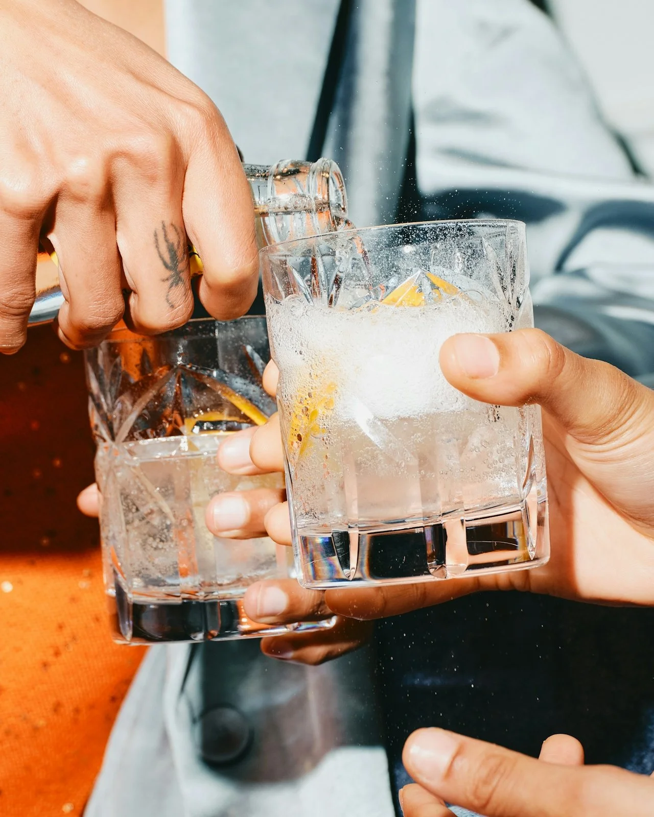 Two people clinking glasses filled with clear liquid and ice, with lemon slices, during daytime.