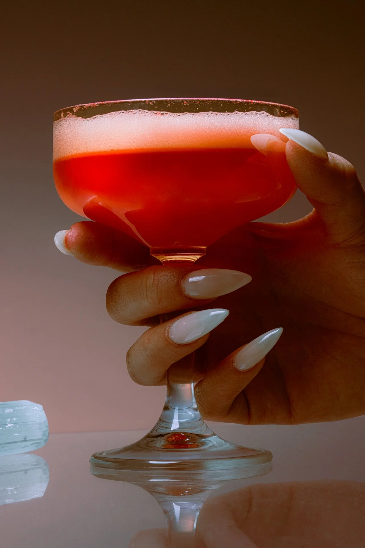 A person holding a pink cocktail glass with a creamy foam on top, set against a neutral background.