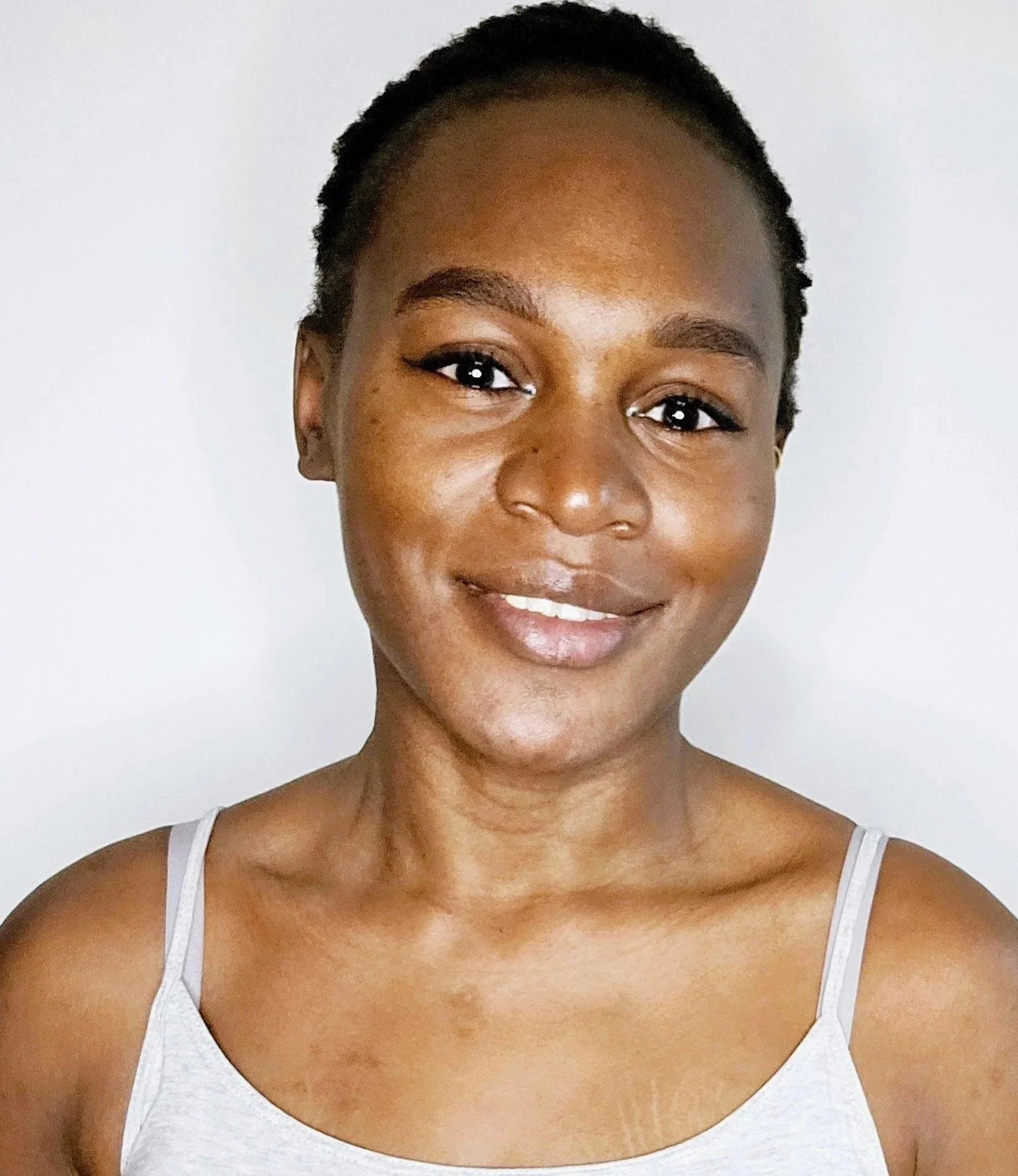 Portrait d'une femme noire avec un sourire, portant un t-shirt gris à fines bretelles, fond blanc.